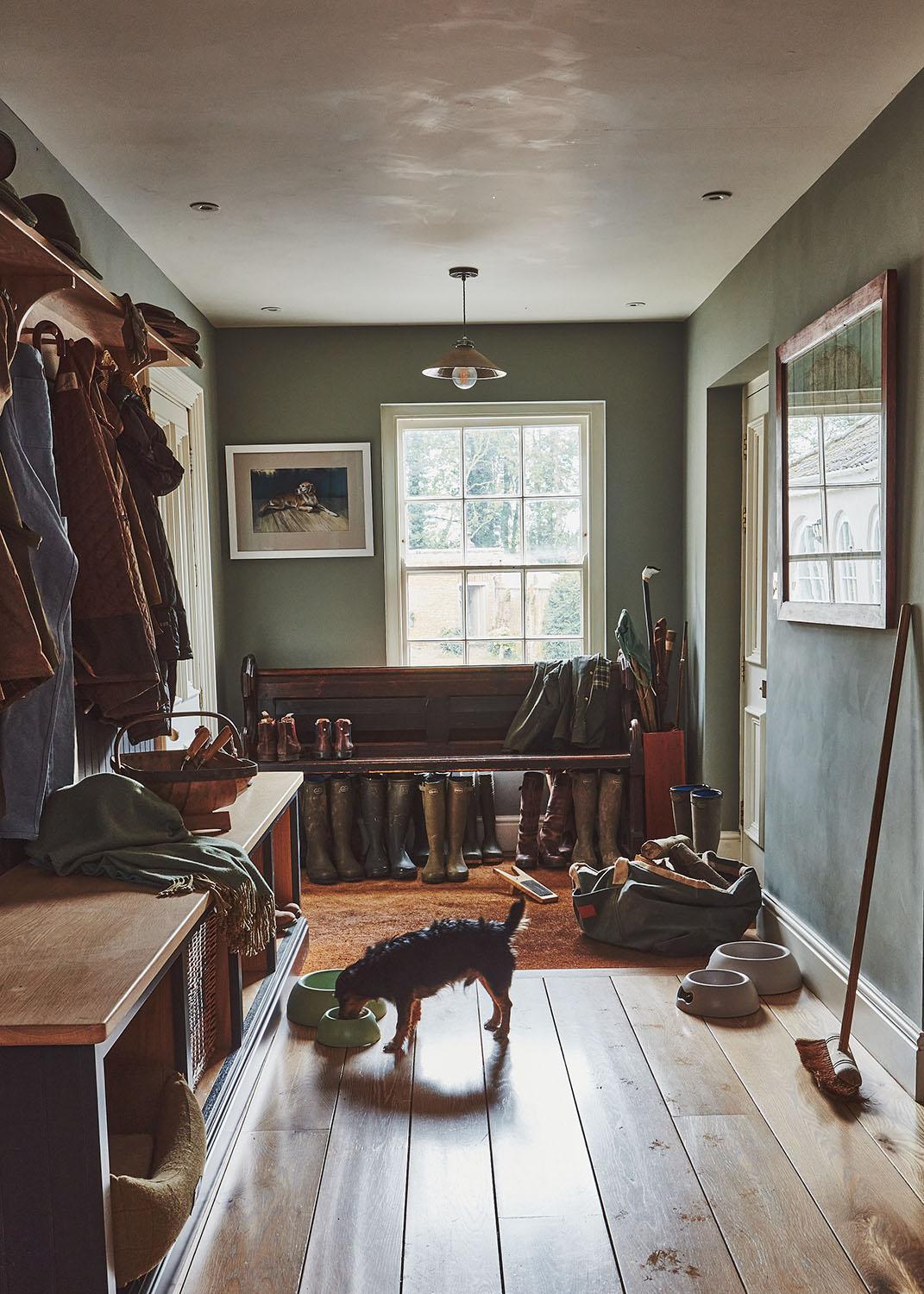 A cozy boot room with wooden floors, benches, coats, boots, and outdoor gear. A small dog eats from a bowl near pet supplies. Sunlight streams through a window at the end of the hallway.