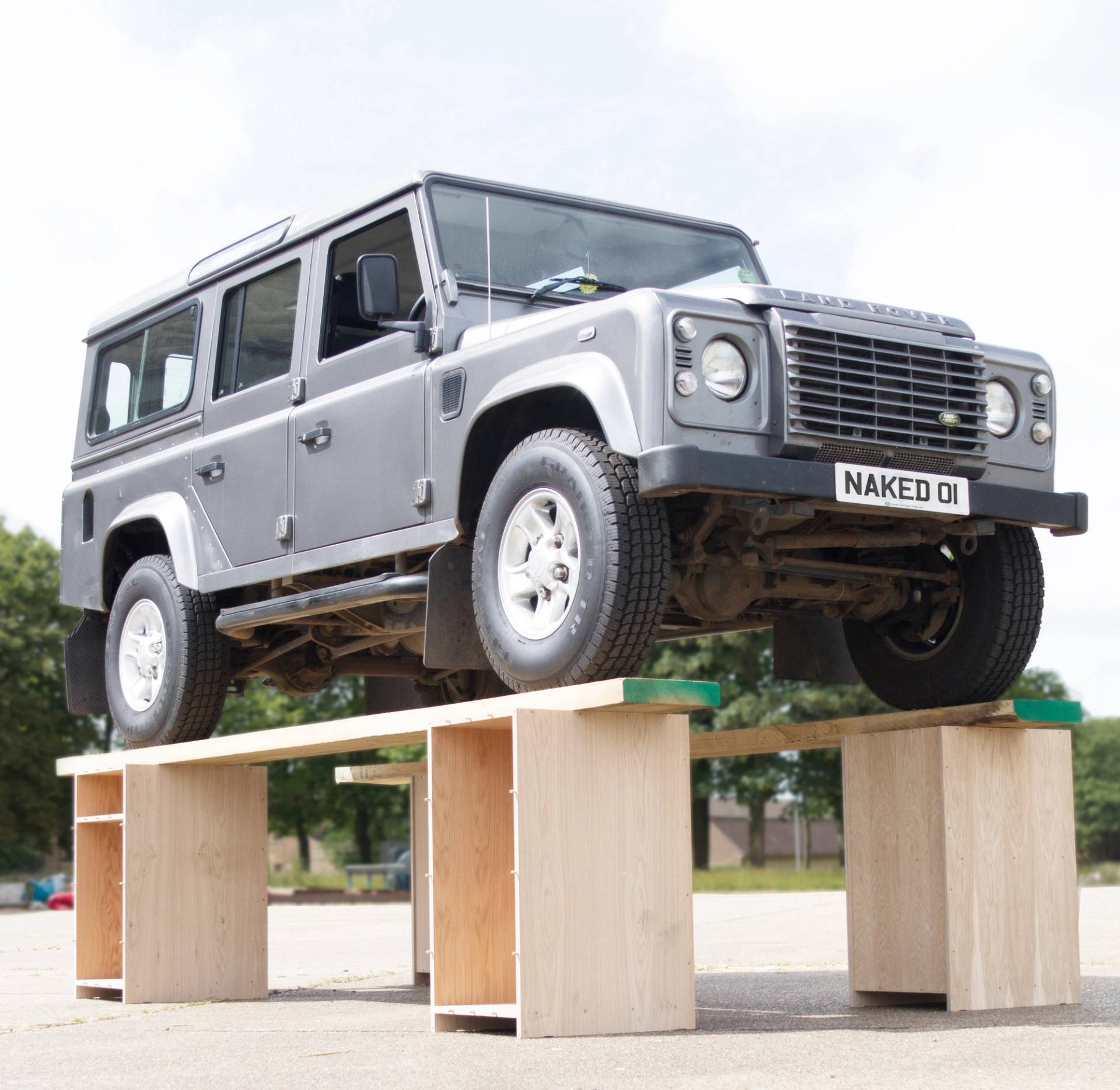 A silver 4x4 with a 'NAKED 01' license plate is elevated on a makeshift wooden platform, showcasing its undercarriage. The background features a cloudy sky and trees.