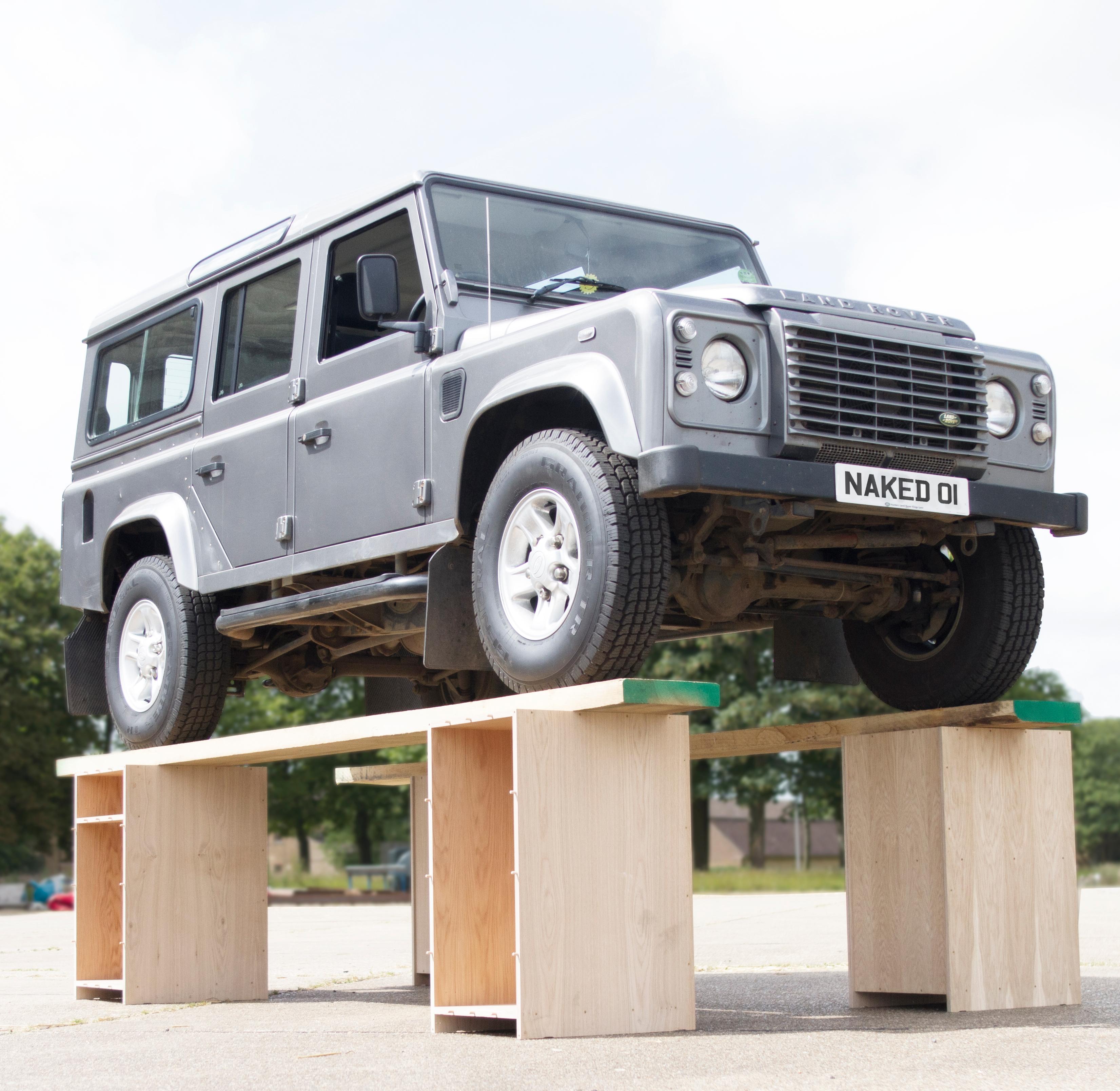 A silver 4x4 with a 'NAKED 01' license plate is elevated on a makeshift wooden platform, showcasing its undercarriage. The background features a cloudy sky and trees.