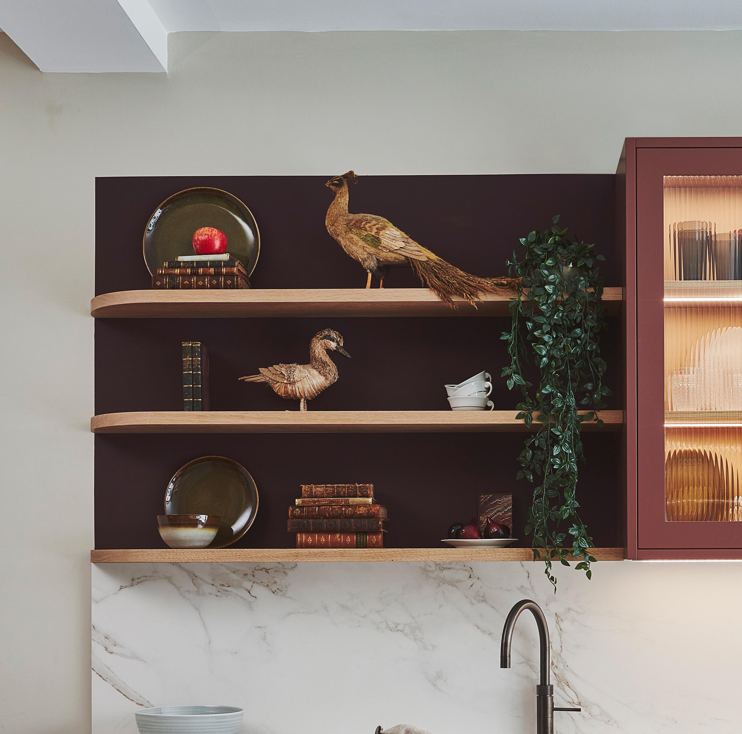 Wooden shelves on a maroon wall display various ceramic items: a tall abstract vase, a pitcher, two wooden cups, a stack of smooth stones, and two small bowls. A row of translucent tile pieces lines the bottom shelf.