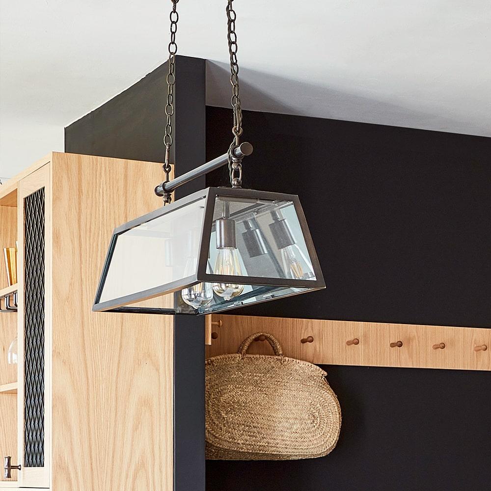 A modern rectangular glass pendant light with three exposed bulbs hangs from the ceiling by two chains above a wooden wall with coat hooks and a woven basket hanging on one hook.