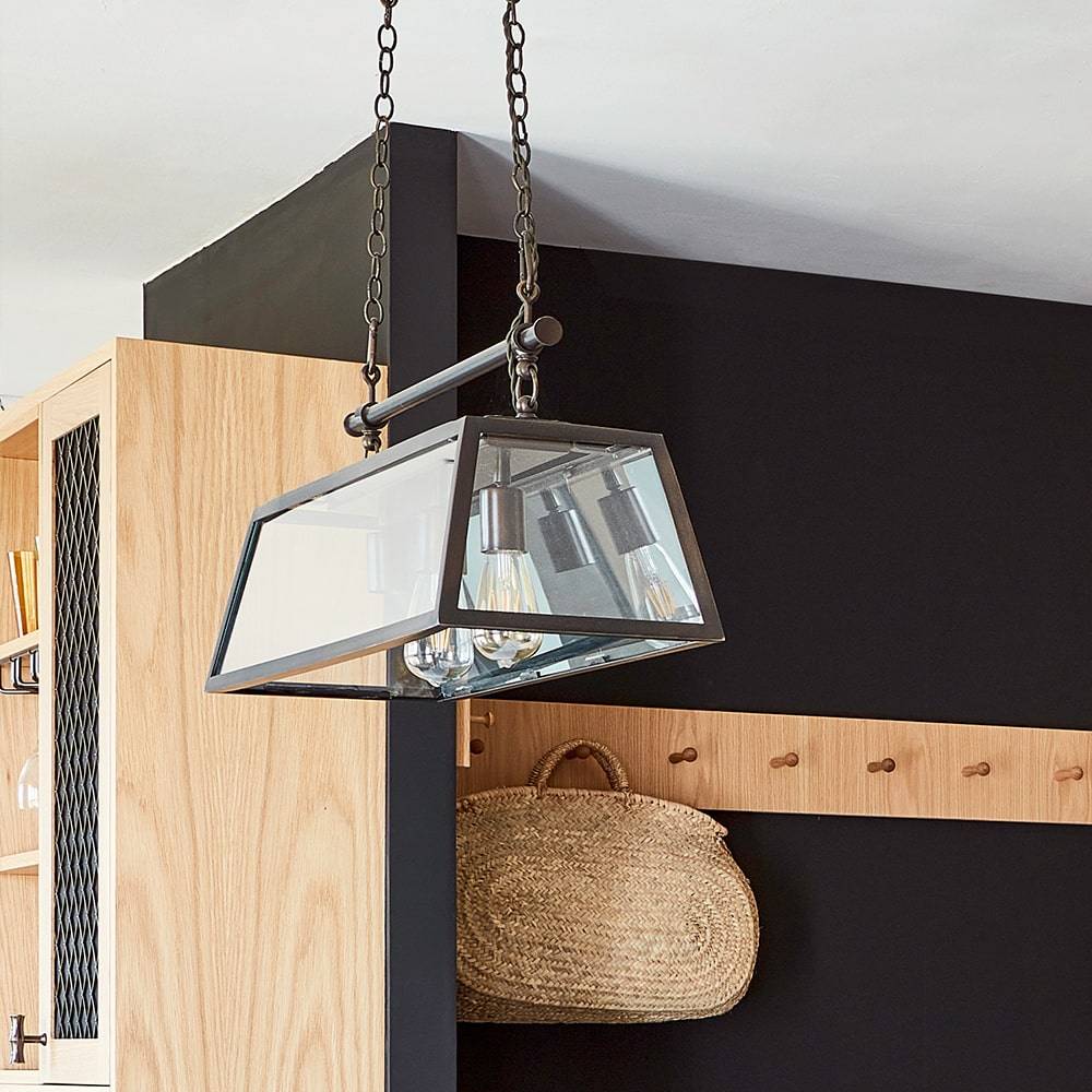 A modern rectangular glass pendant light with three exposed bulbs hangs from the ceiling by two chains above a wooden wall with coat hooks and a woven basket hanging on one hook.
