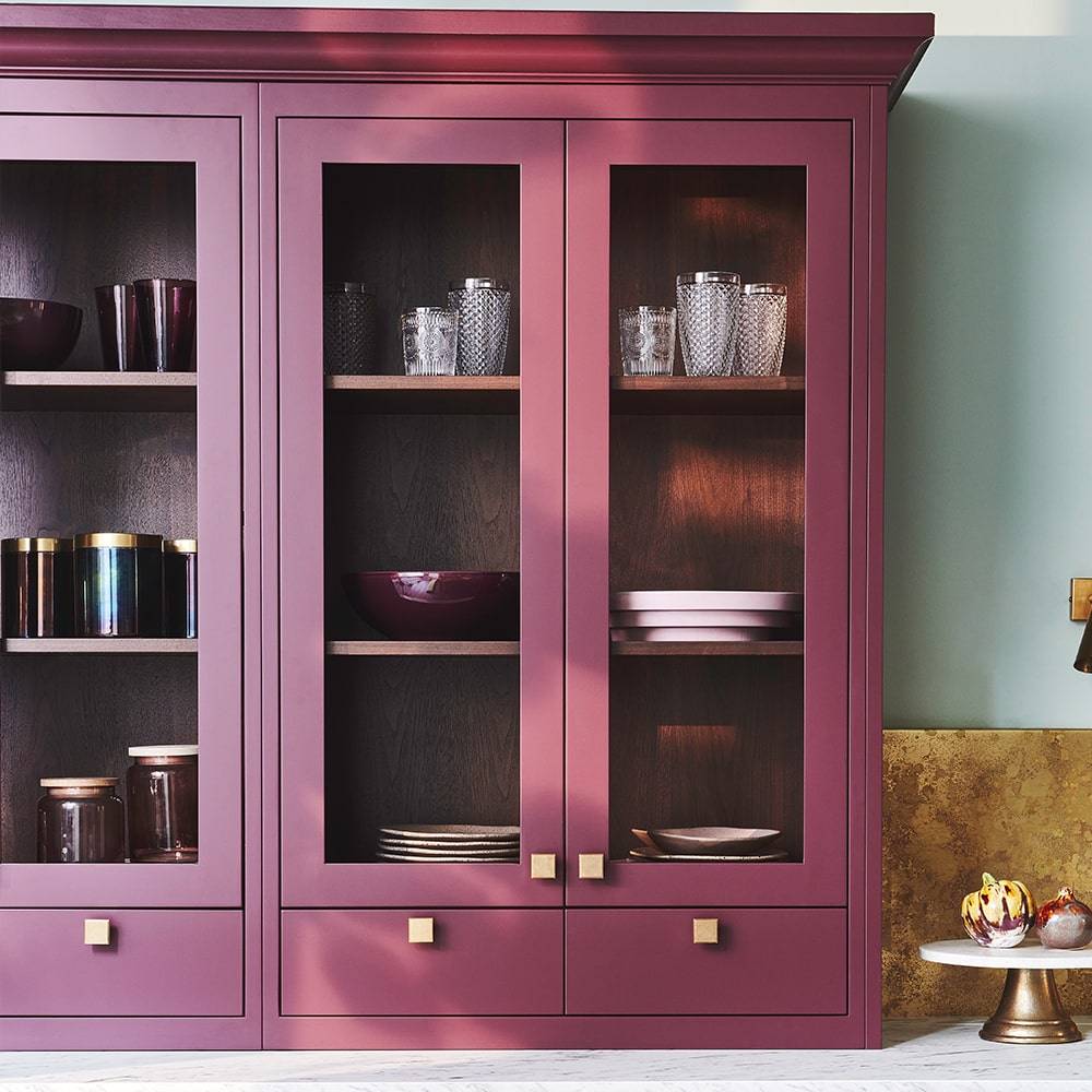 A burgundy kitchen cabinet with glass doors displays neatly arranged glassware, bowls, and plates. The cabinet has gold knobs and is set against a light teal and gold wall. A cake stand is visible on the side.