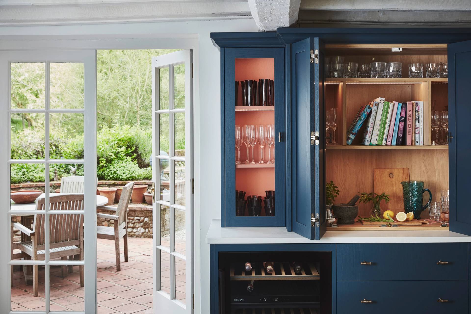 A blue kitchen cabinet with open doors reveals glassware, cookbooks, and a cutting board with a knife and sliced lemon. French doors lead out to a patio with wooden chairs and a table, surrounded by greenery.