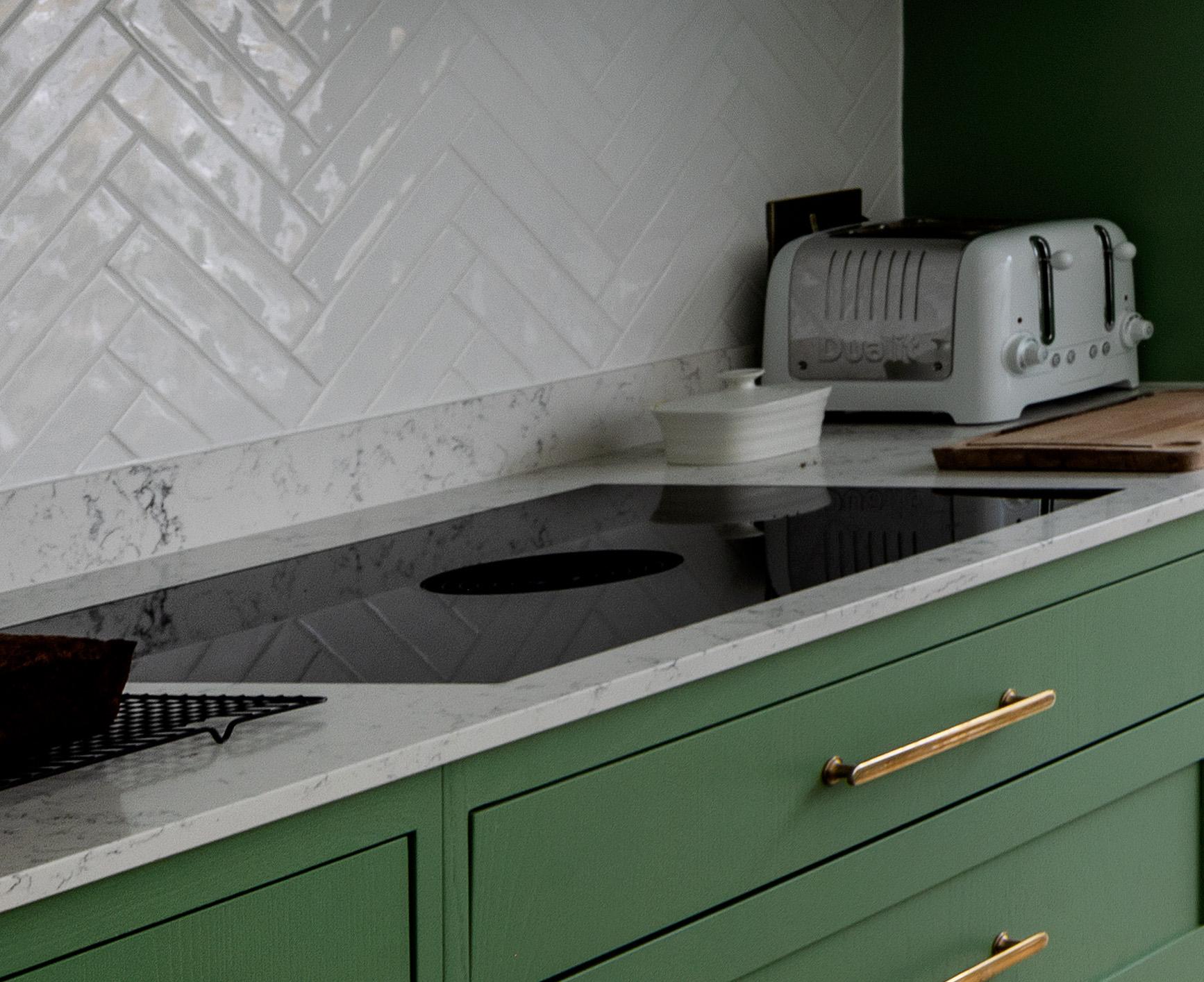 A modern kitchen counter with green cabinets, gold handles, a white marble countertop, a black electric cooktop, a white herringbone tile backsplash, a gray toaster, and a small white container.