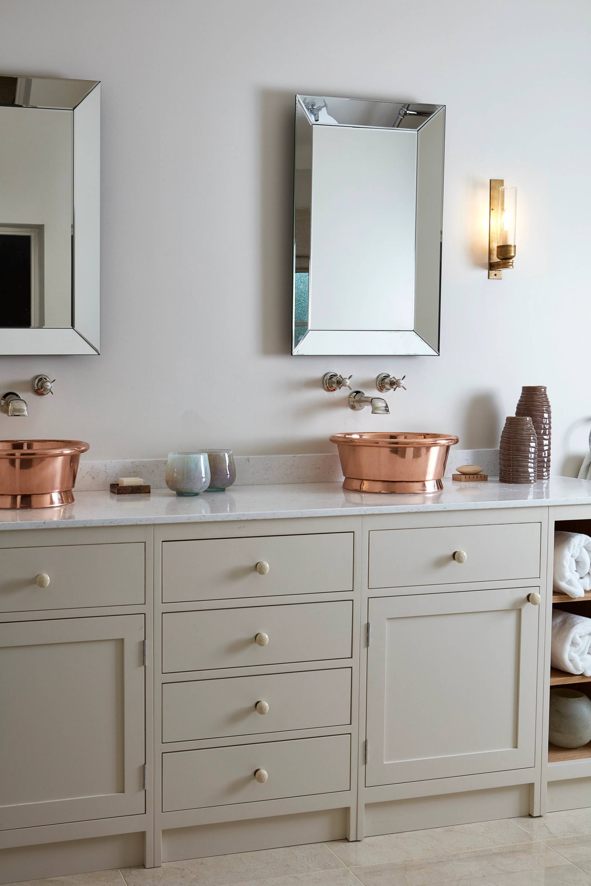 Modern bathroom with dual sinks in copper basins on a beige countertop. Two rectangular mirrors hang above the sinks. Towels are neatly stacked on open shelves to the right, next to a decorative vase and soap dishes. A wall sconce provides lighting.