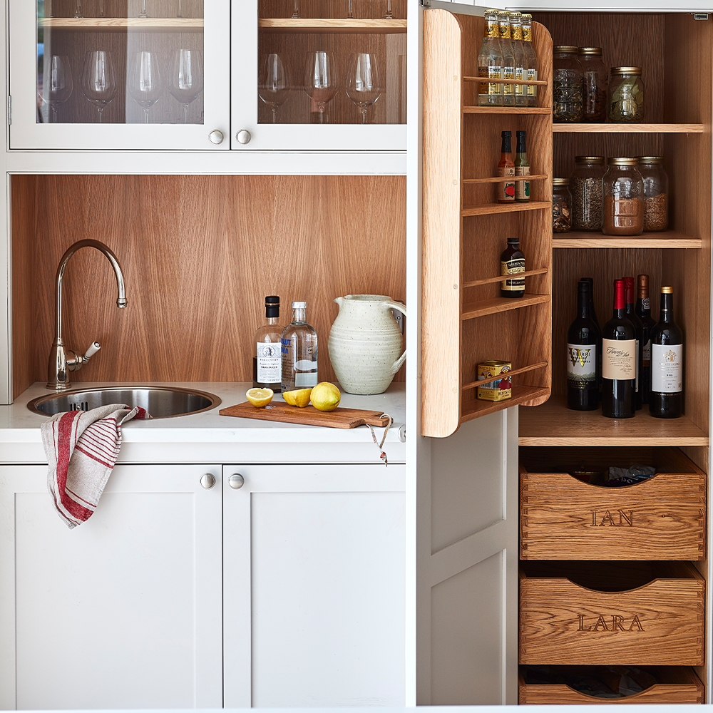 A small kitchen nook with a sink, bottles, a chopping board with lemons, and a pitcher. Next to it, an open cabinet holds spices, jars, wine bottles, and wooden drawers labeled IAN and LARA.