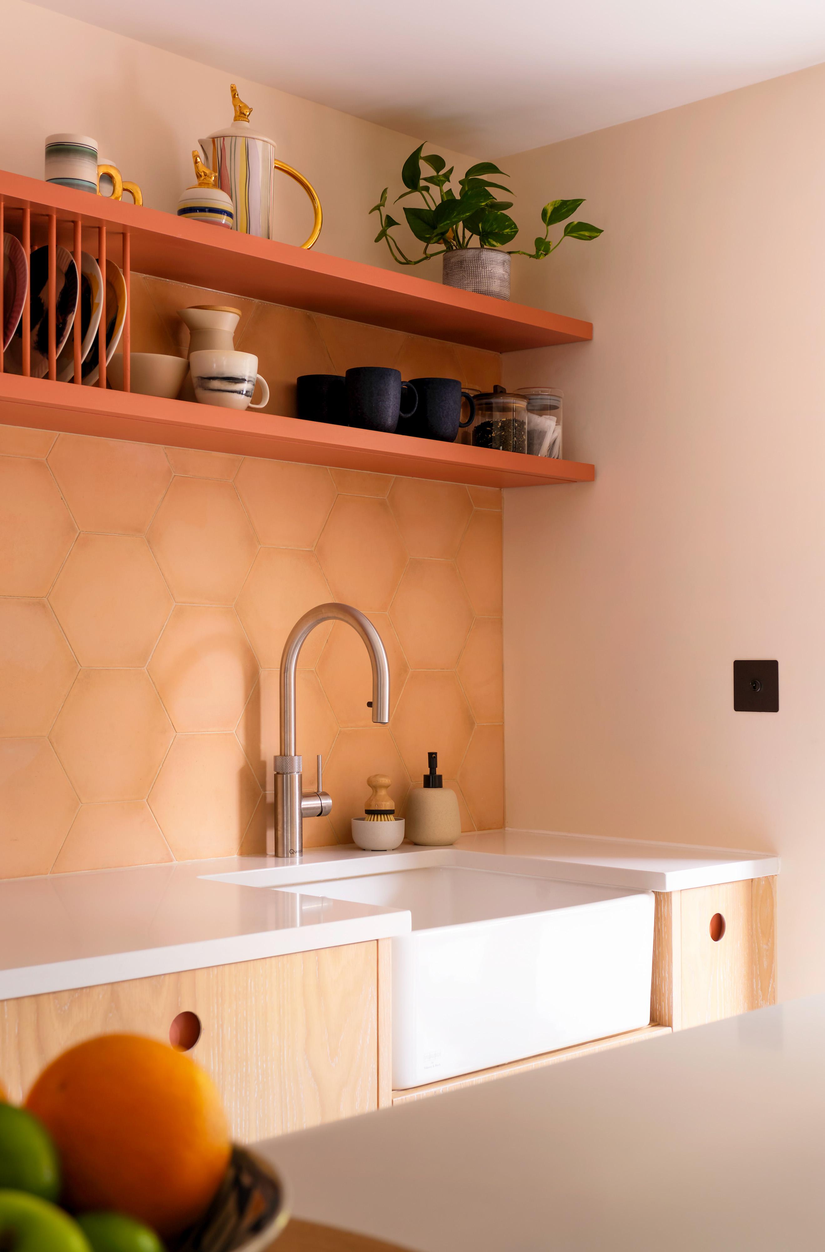Modern kitchen with hexagonal peach-colored tiles, a white sink with a silver faucet, and wooden shelves holding cups and plants. A bowl of fruit is slightly visible in the foreground