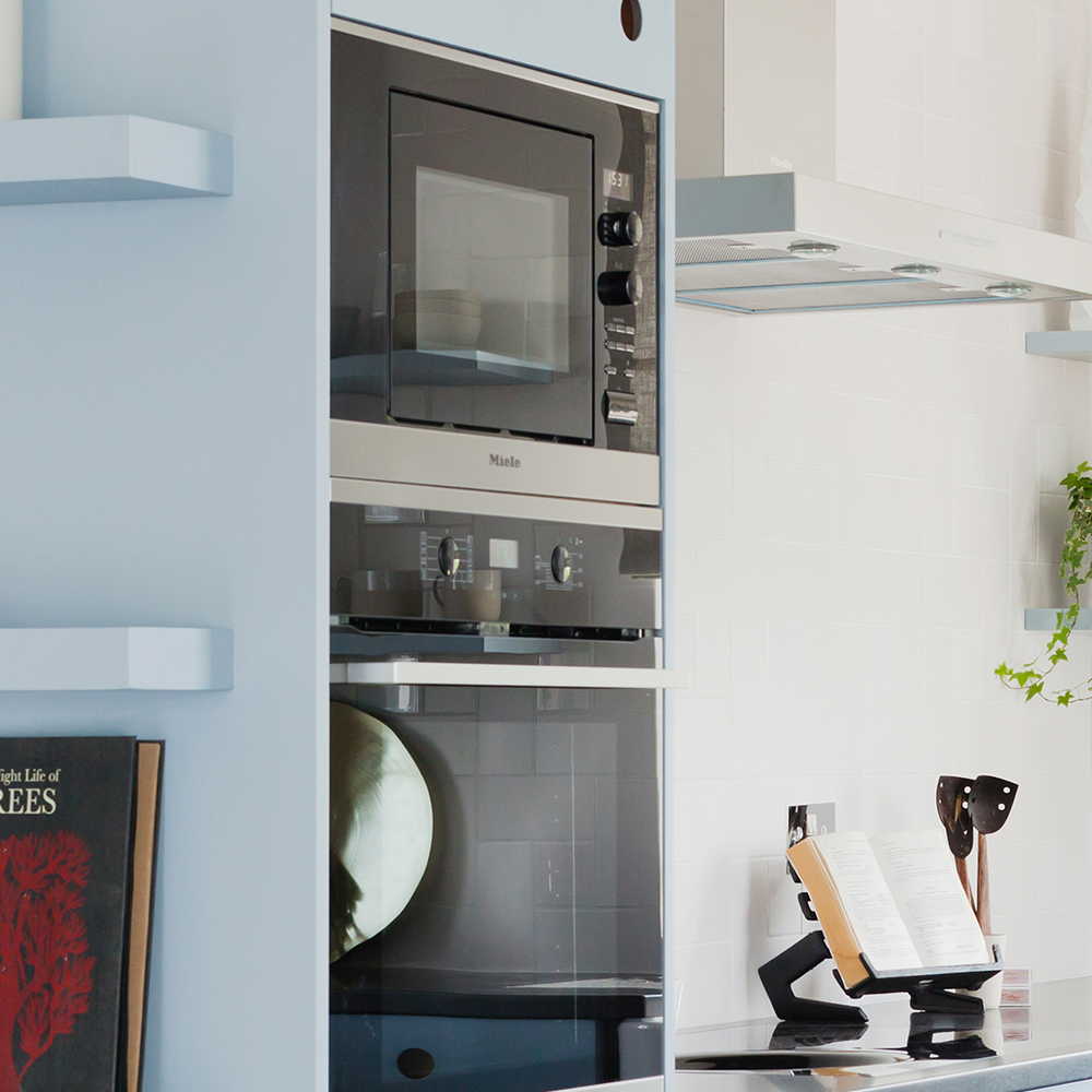 A modern kitchen with a built-in microwave and oven, an open cookbook on a stand, utensils in a holder, and a potted plant on a white countertop next to a range hood.