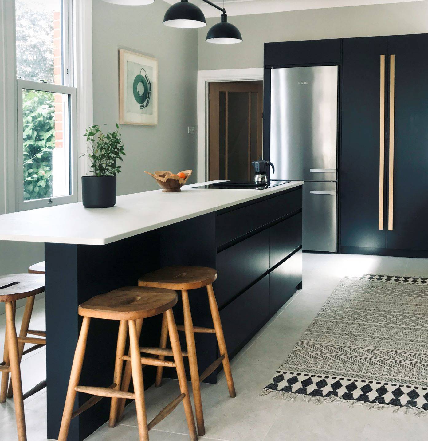Modern kitchen with a sleek black island counter featuring wooden stools. A stainless steel fridge stands nearby, complemented by black cabinets with wooden handles. Soft lighting from hanging lamps and a patterned rug add warmth to the space.