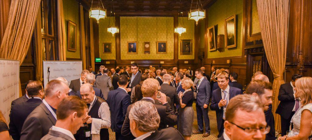 A crowded, formal evening reception taking place in a grand, wood-paneled hall featuring gold drapes, portraits, and hanging light fixtures.