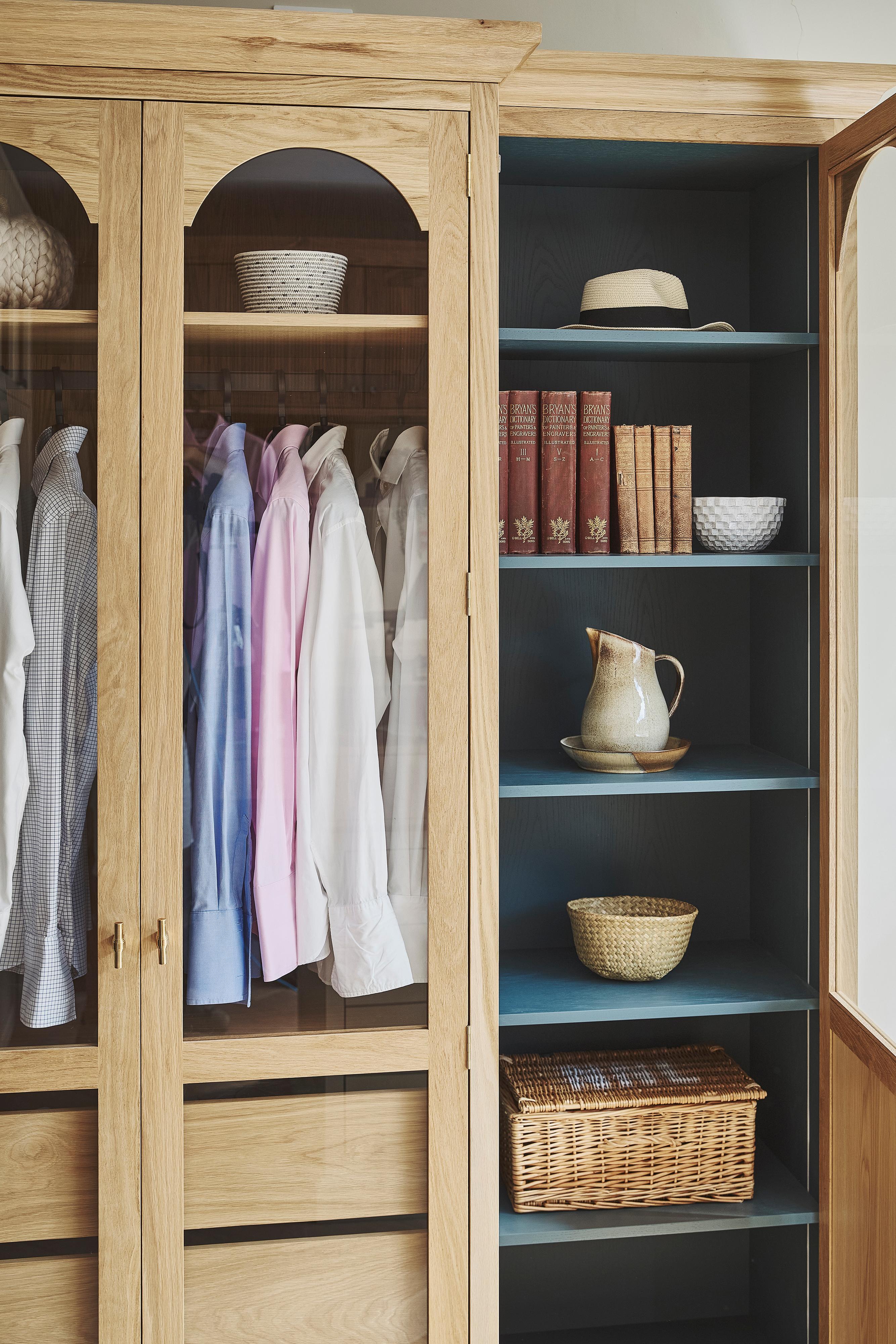 An oak wardrobe with a glass door is partially open, revealing three wooden drawers and several shirts hanging above them.
