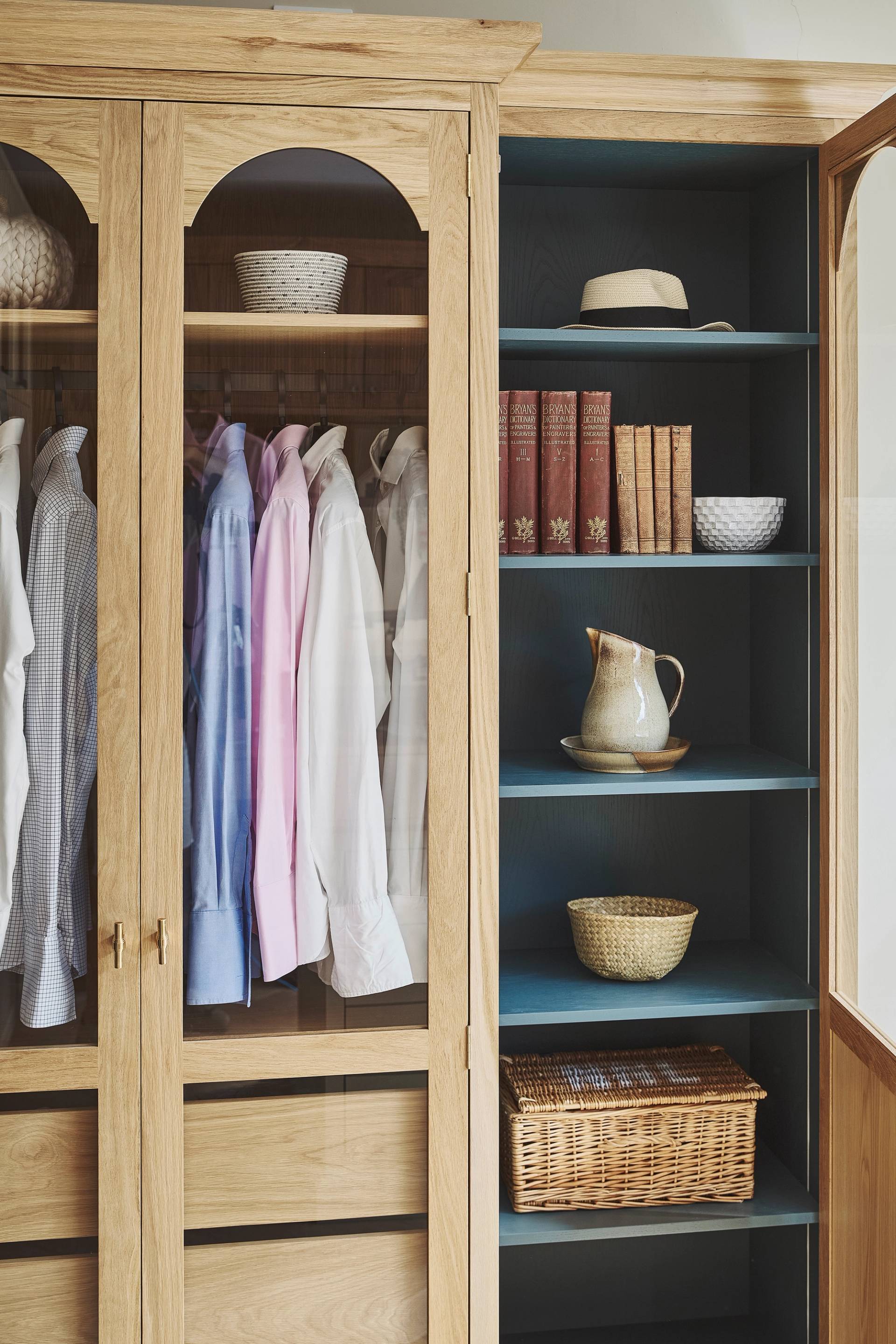 An oak wardrobe with a glass door is partially open, revealing three wooden drawers and several shirts hanging above them.