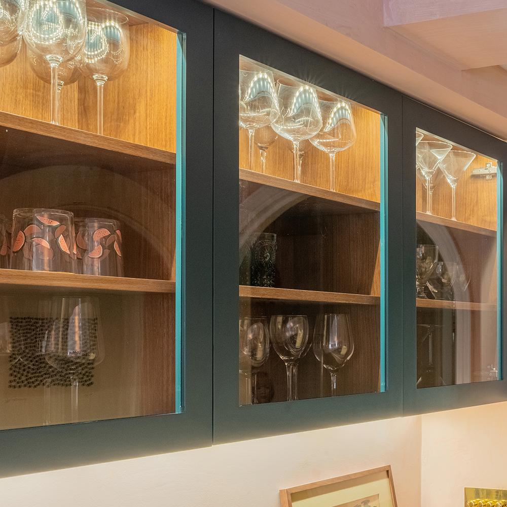 Glass-front cabinets with wooden shelves displaying wine glasses, cocktail glasses, and other glassware, all neatly arranged and lit by built-in lighting.