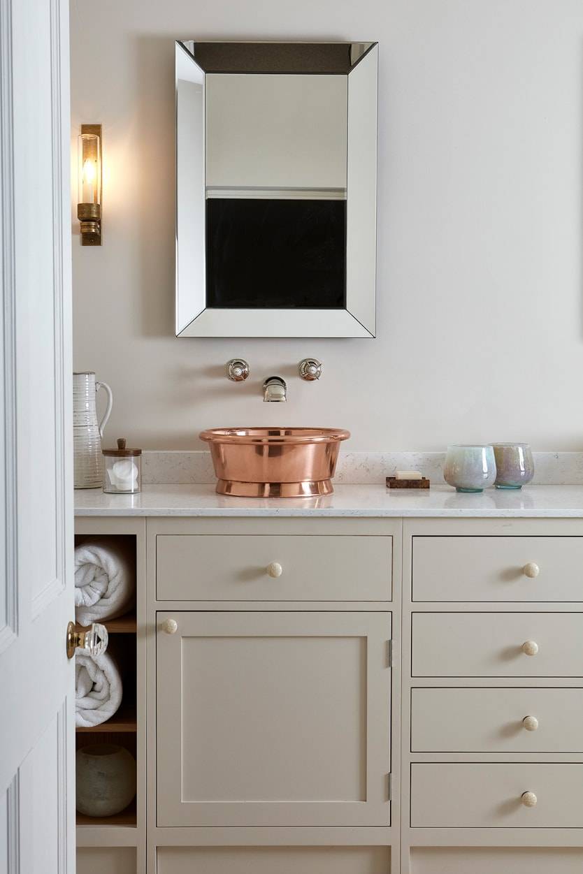 A modern bathroom with cream cabinets, a countertop holding a shiny copper basin sink, a wall-mounted faucet, a square mirror, a wall sconce, and shelves with rolled white towels and decorative items.