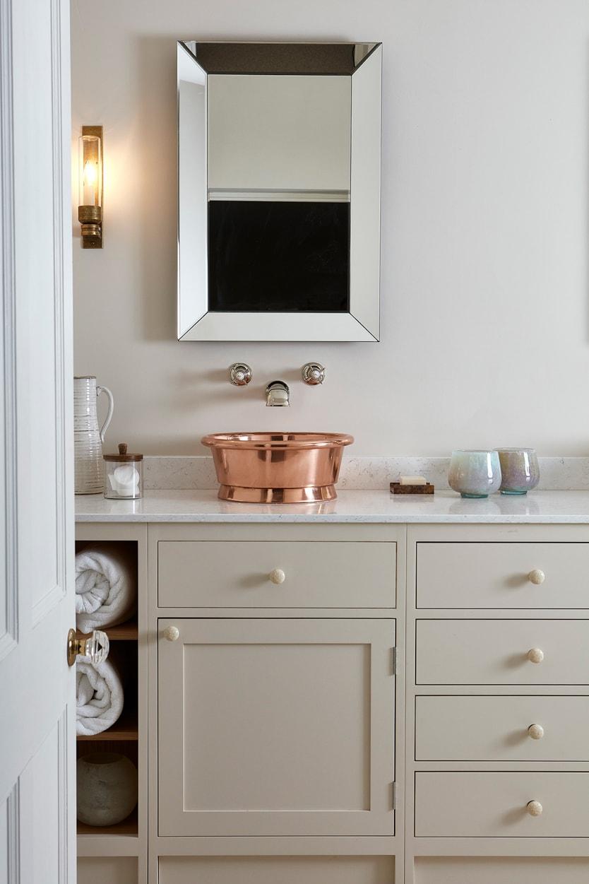 A modern bathroom with cream cabinets, a countertop holding a shiny copper basin sink, a wall-mounted faucet, a square mirror, a wall sconce, and shelves with rolled white towels and decorative items.