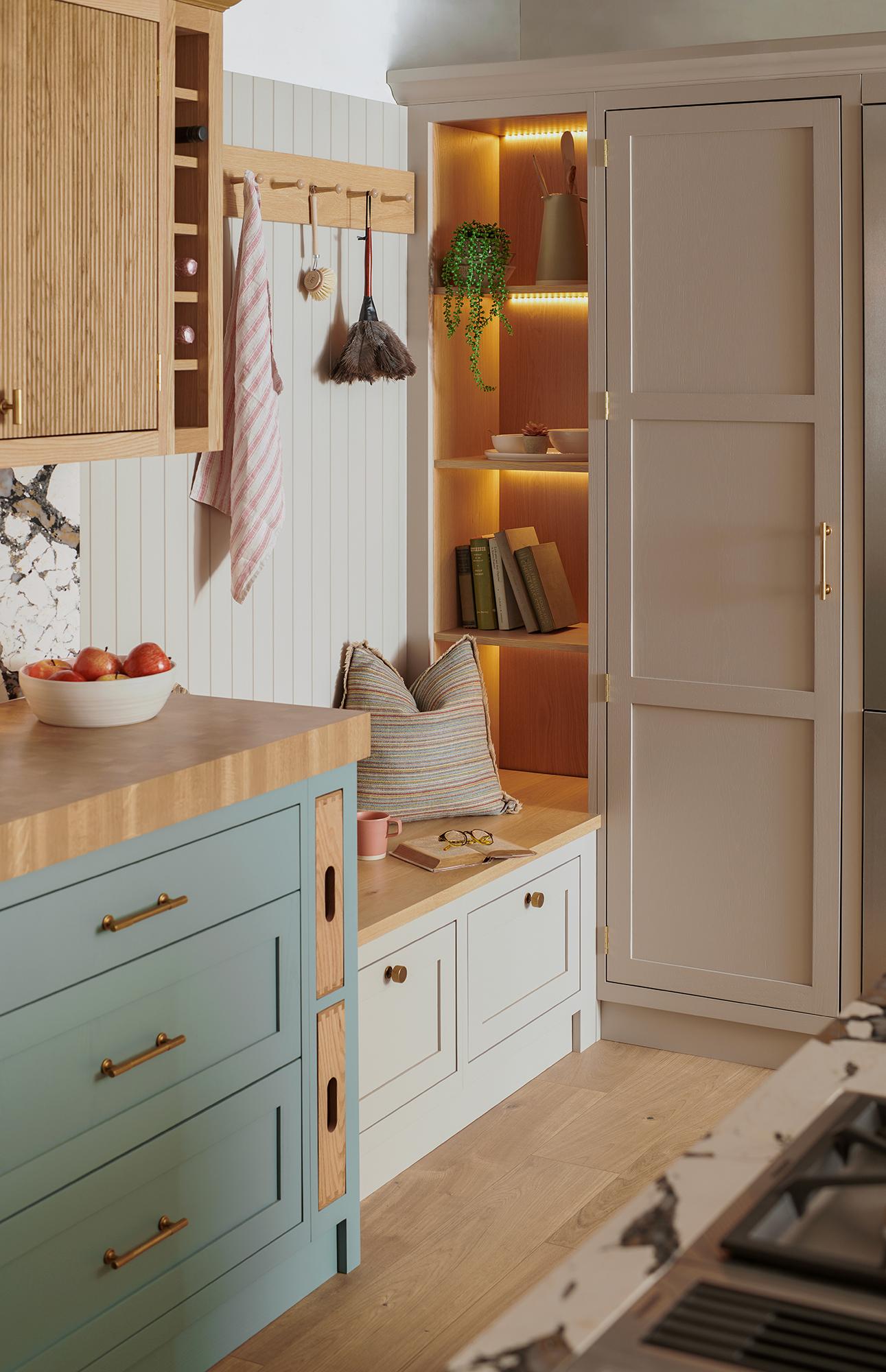 A cozy kitchen corner featuring a built-in reading nook with shelves holding books and a plant. The nook is illuminated by warm LED lights and has a cushioned bench. The kitchen has light blue and wooden cabinets, a bowl of tomatoes, and stylish decor.