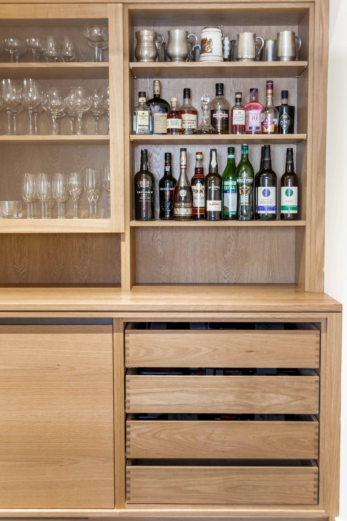 A wooden cabinet with shelves displaying various glassware on the left and bottles of liquor and metal mugs on the right. The lower section has closed wooden drawers and panels.