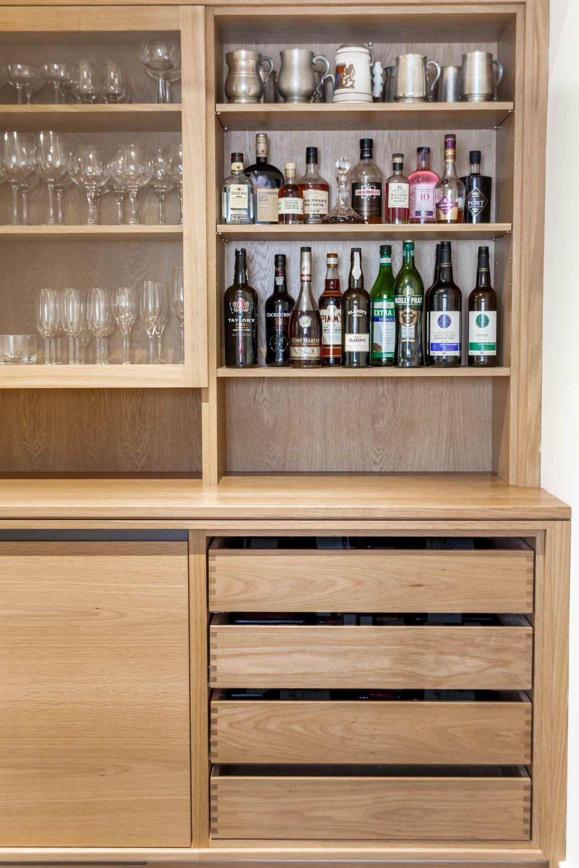 A wooden cabinet with shelves displaying various glassware on the left and bottles of liquor and metal mugs on the right. The lower section has closed wooden drawers and panels.