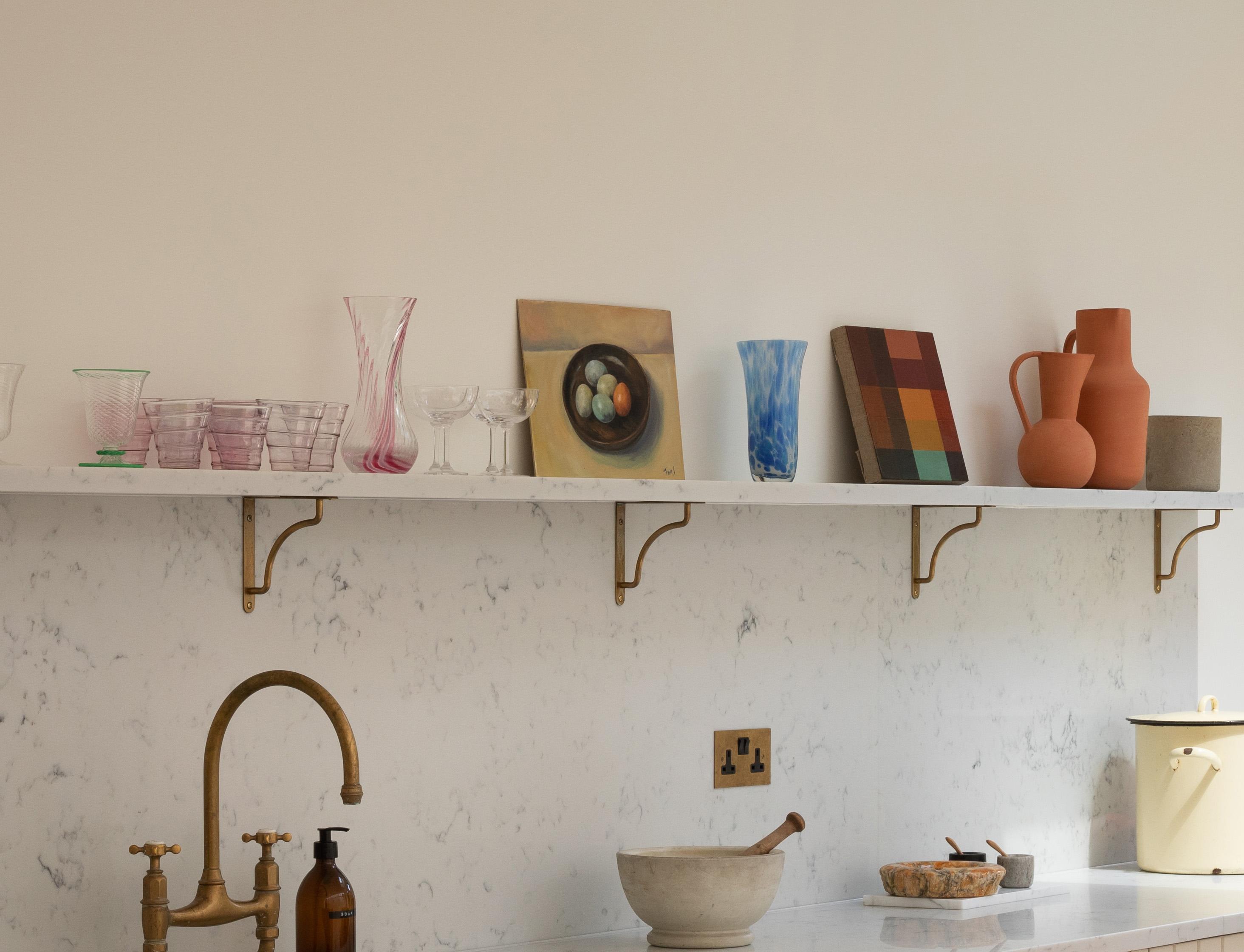 A kitchen shelf displays glassware, vases, framed art, and ceramics above a marble countertop with a brass faucet, a mortar and pestle, and a pale yellow container.