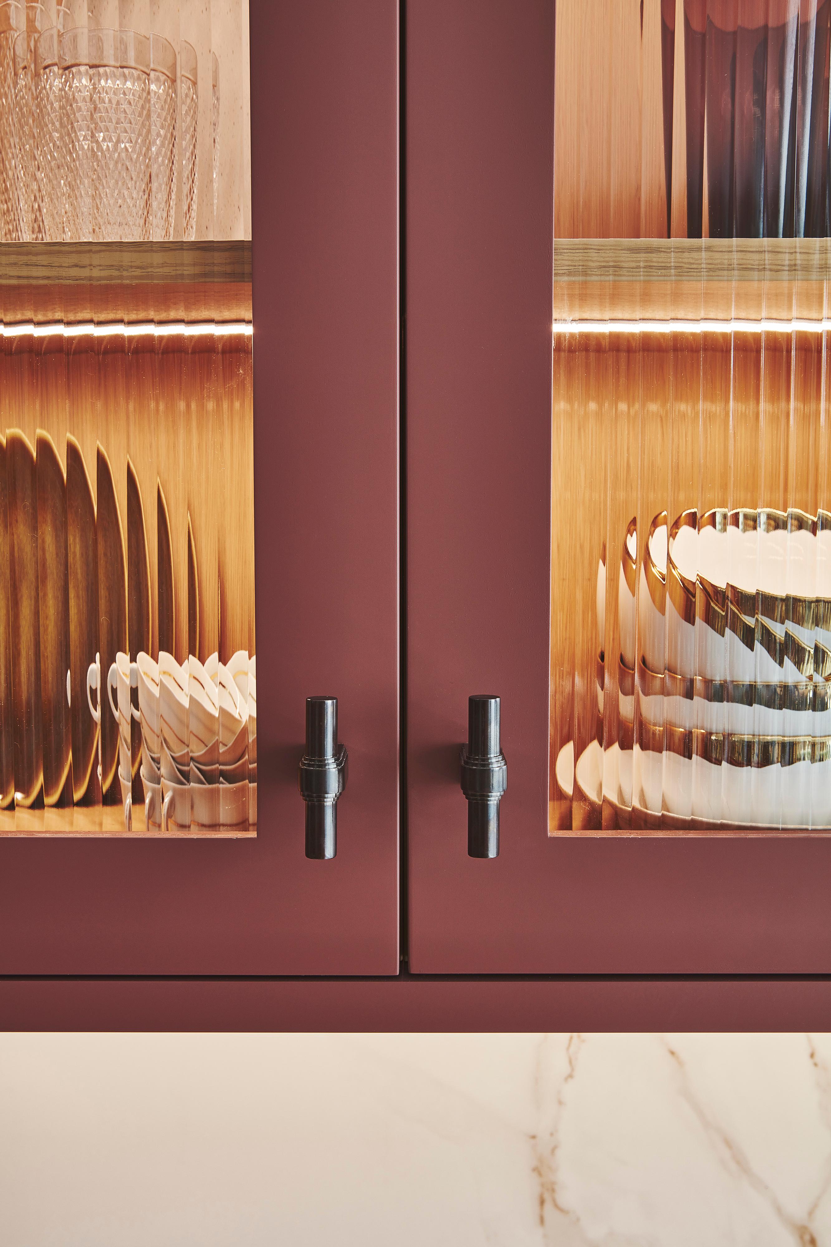 Close-up of a kitchen cabinet with textured glass doors and black handles. Inside are glassware and stacked plates, softly illuminated by warm lighting, on a marble countertop below.