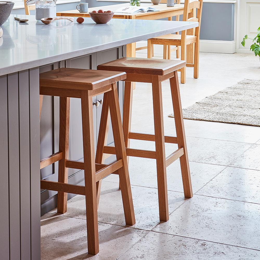 Two wooden bar stools are placed beside a kitchen island with a light countertop in a bright, modern kitchen. The room features light flooring, natural light, and breakfast items on the counter.
