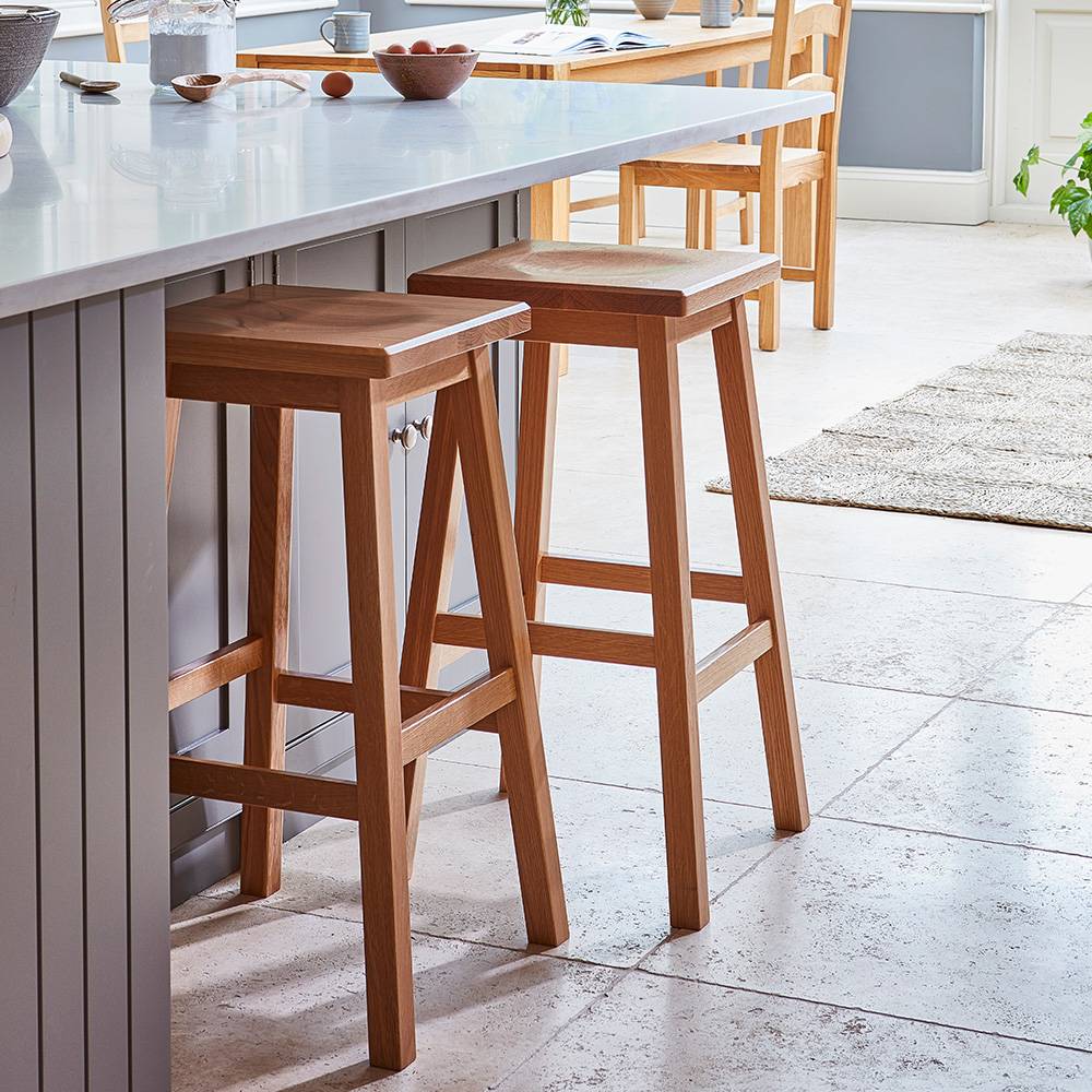 Two wooden bar stools are placed beside a kitchen island with a light countertop in a bright, modern kitchen. The room features light flooring, natural light, and breakfast items on the counter.