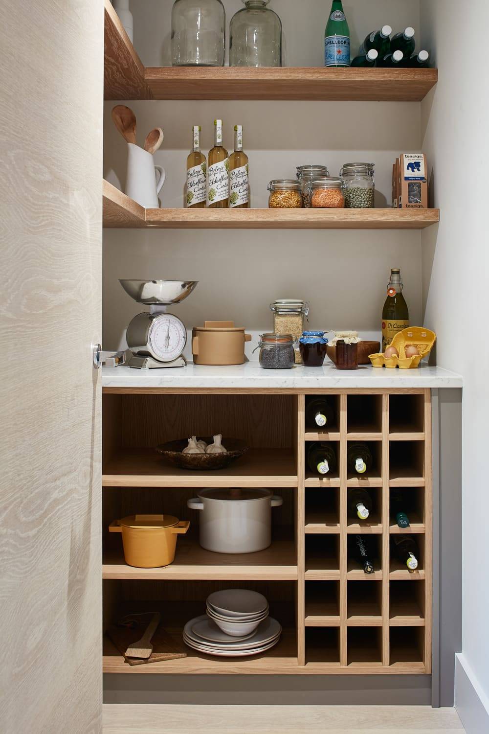 A well-organized pantry with shelves holding jars, bottles, canned goods, glass containers, and kitchen tools. Below are cubbies storing wine bottles, pots, dishes, and a kitchen scale on the counter.