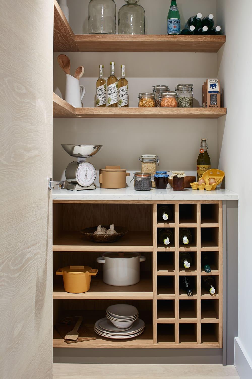 A well-organized pantry with shelves holding jars, bottles, canned goods, glass containers, and kitchen tools. Below are cubbies storing wine bottles, pots, dishes, and a kitchen scale on the counter.