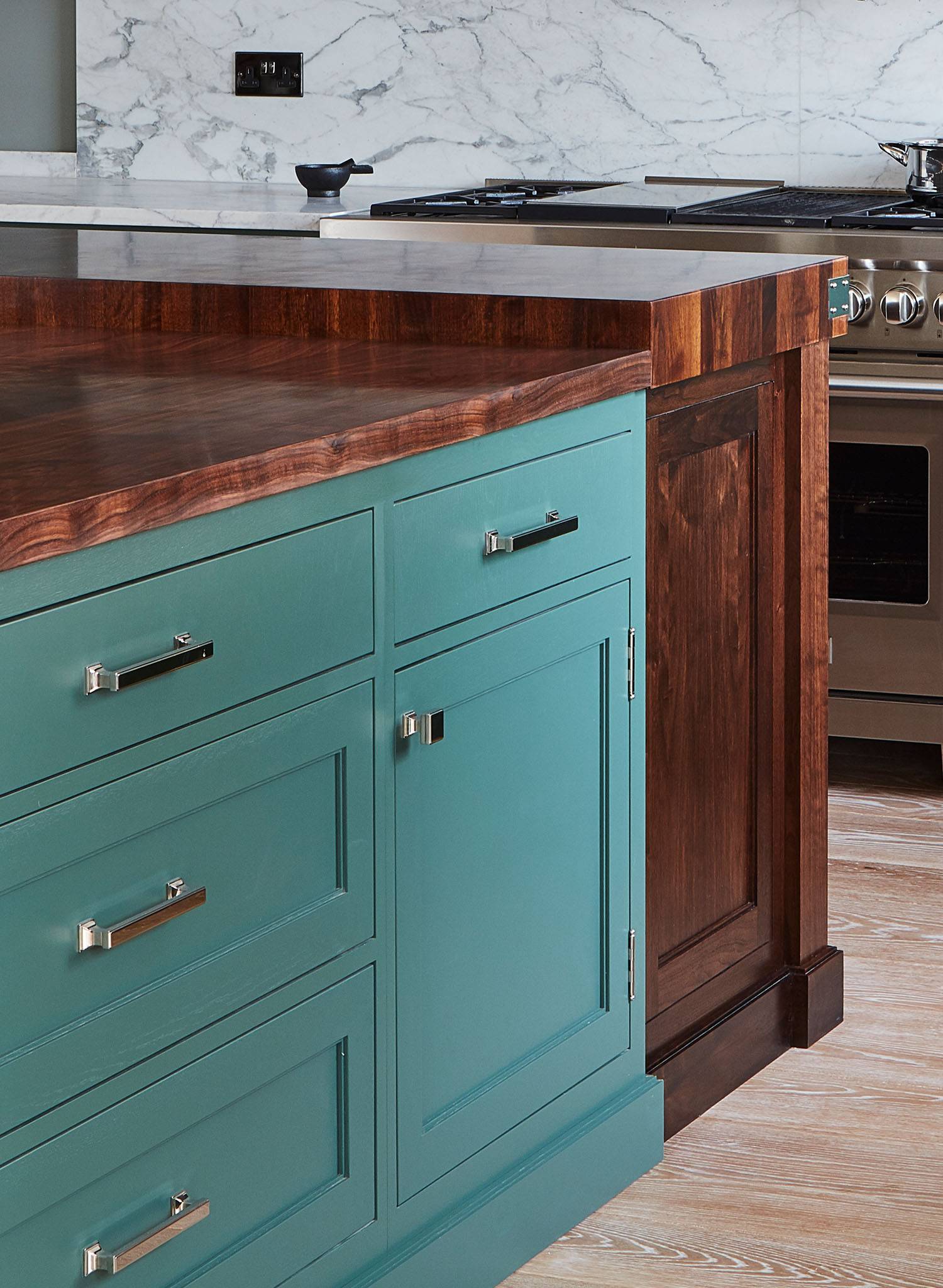 A kitchen island with teal cabinet drawers and silver handles, featuring a dark wood countertop; marble backsplash and stainless steel stove are visible in the background.