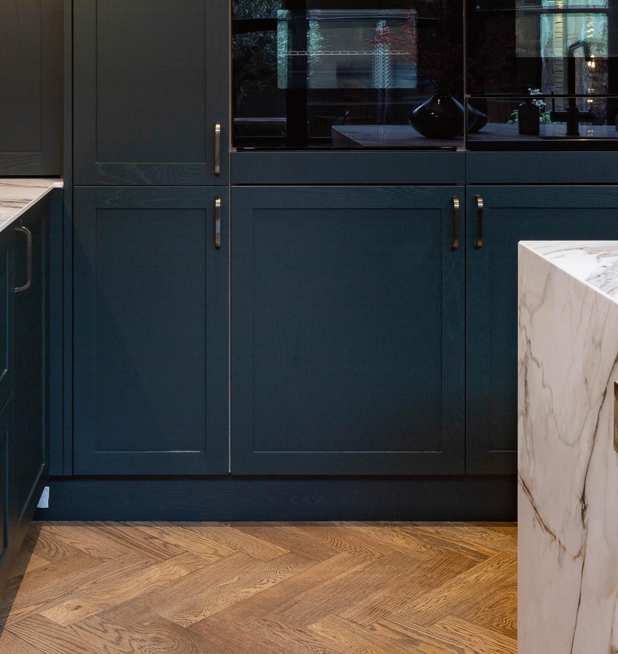 Dark green kitchen cabinets with brass handles are set against a chevron-patterned wooden floor; a white marble countertop is visible on the right side of the image.