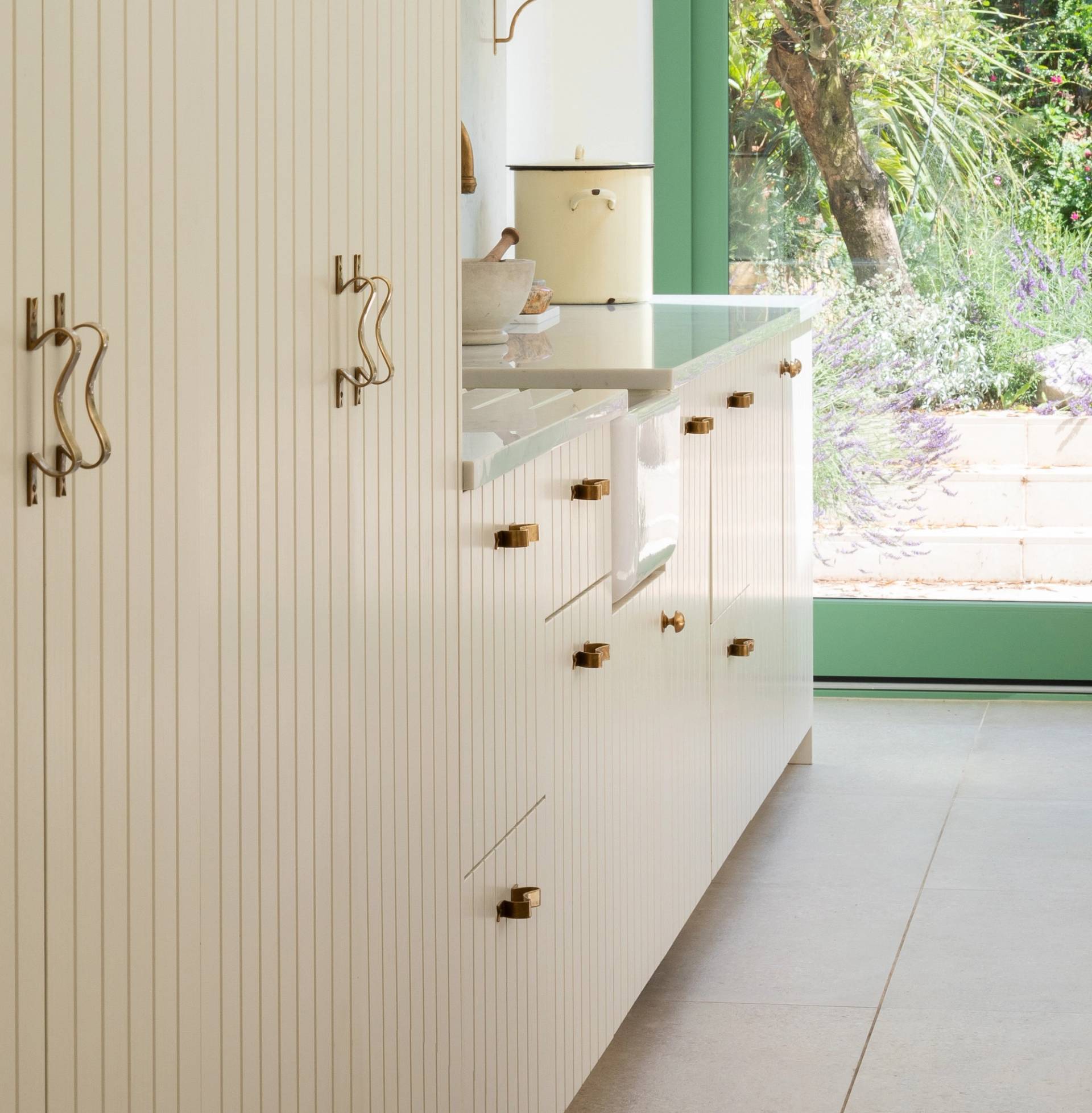 Bright modern kitchen with white cabinets, gold handles, and a large window overlooking a lush green garden. Light pours in, illuminating the minimalist, clean design and natural outdoor view.
