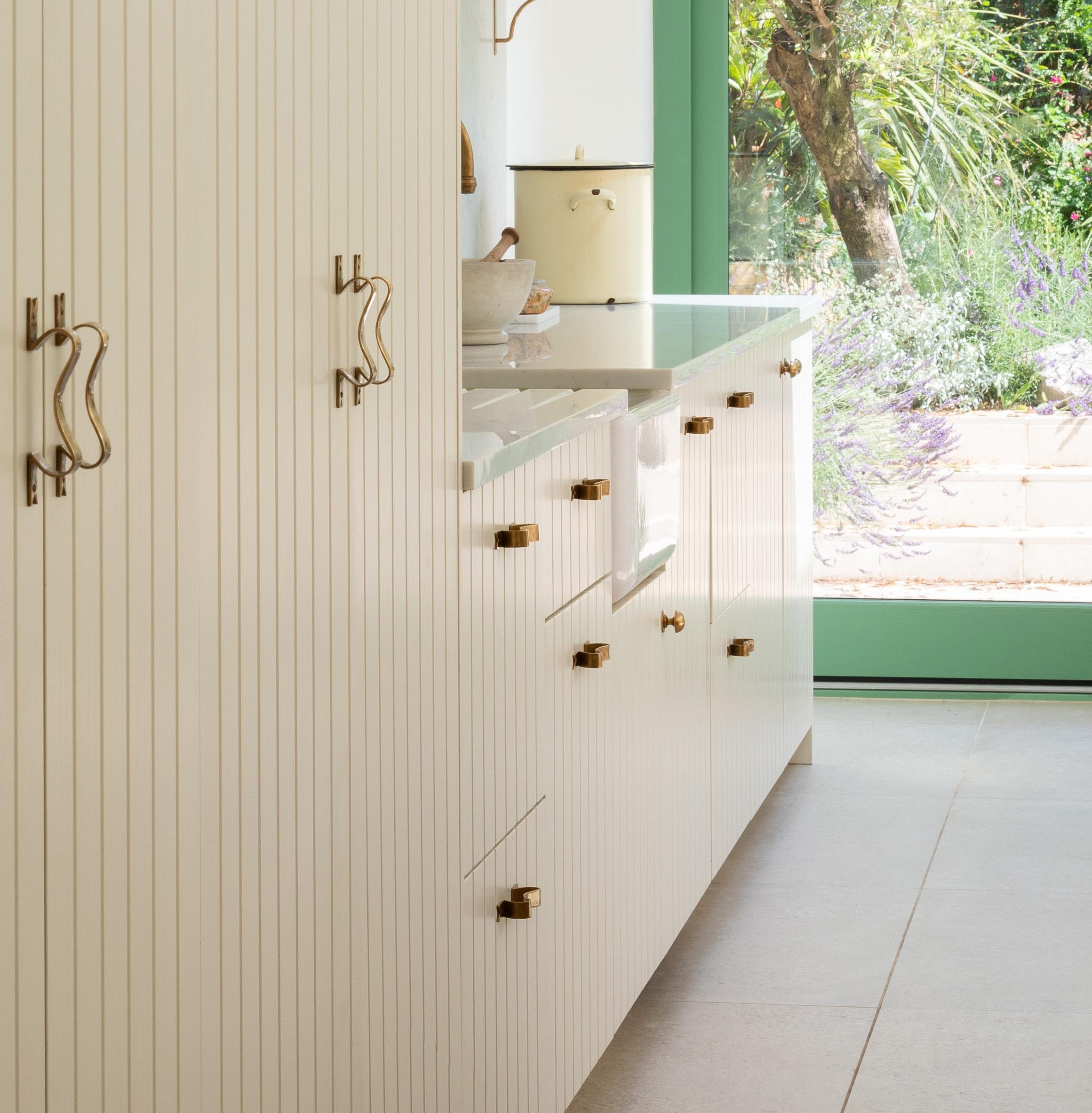 Bright modern kitchen with white cabinets, gold handles, and a large window overlooking a lush green garden. Light pours in, illuminating the minimalist, clean design and natural outdoor view.