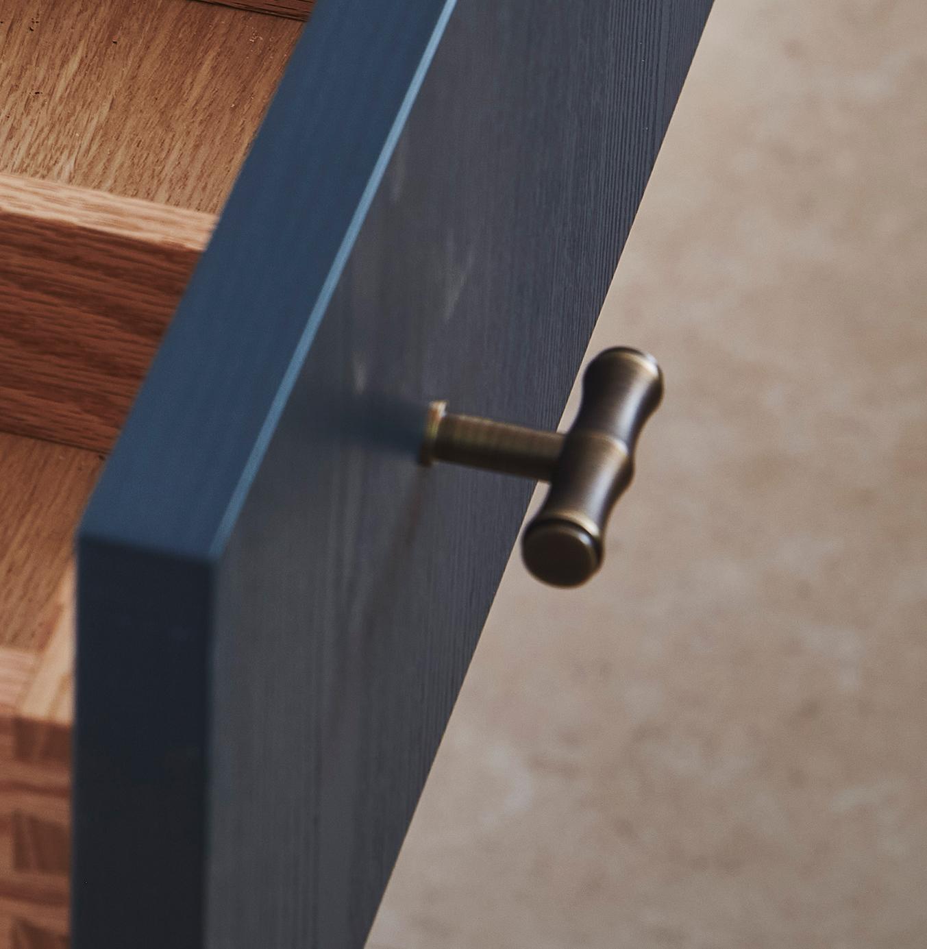 A close-up of a partially open drawer with a dark blue front, a brass T-shaped handle, and light wood interior dividers. The background is a neutral, blurred surface.