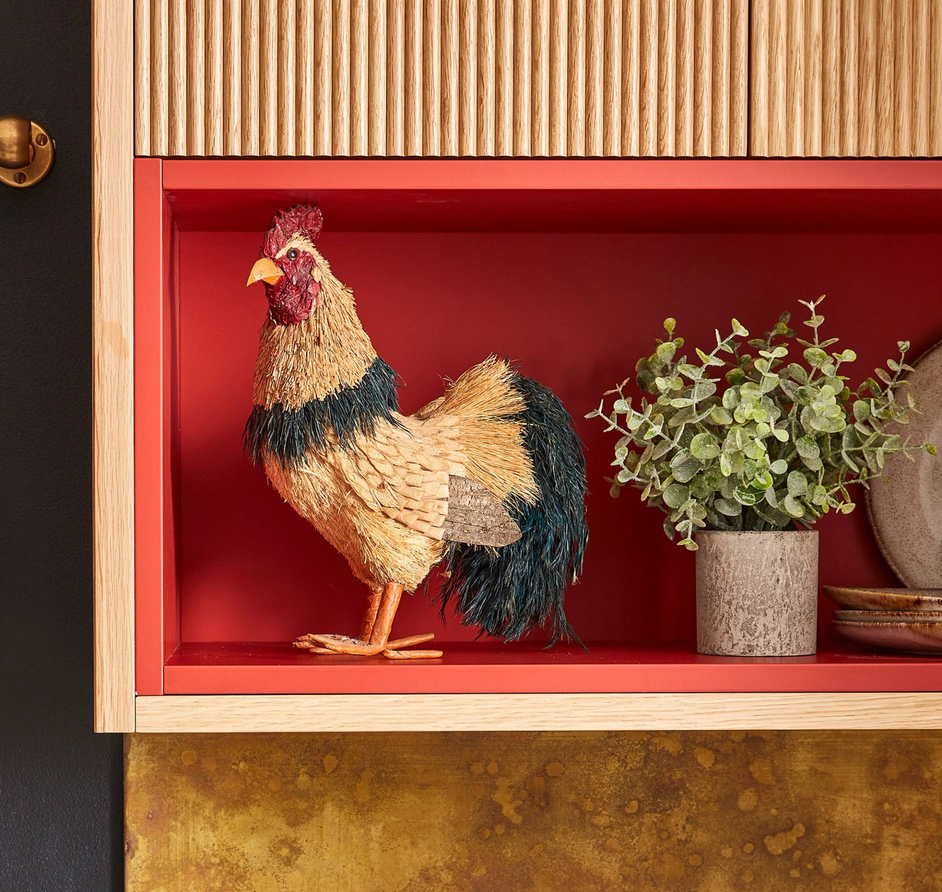 A decorative rooster figurine made of straw stands on a red shelf, next to a potted plant and a plate. The shelf is surrounded by wood and brass accents.