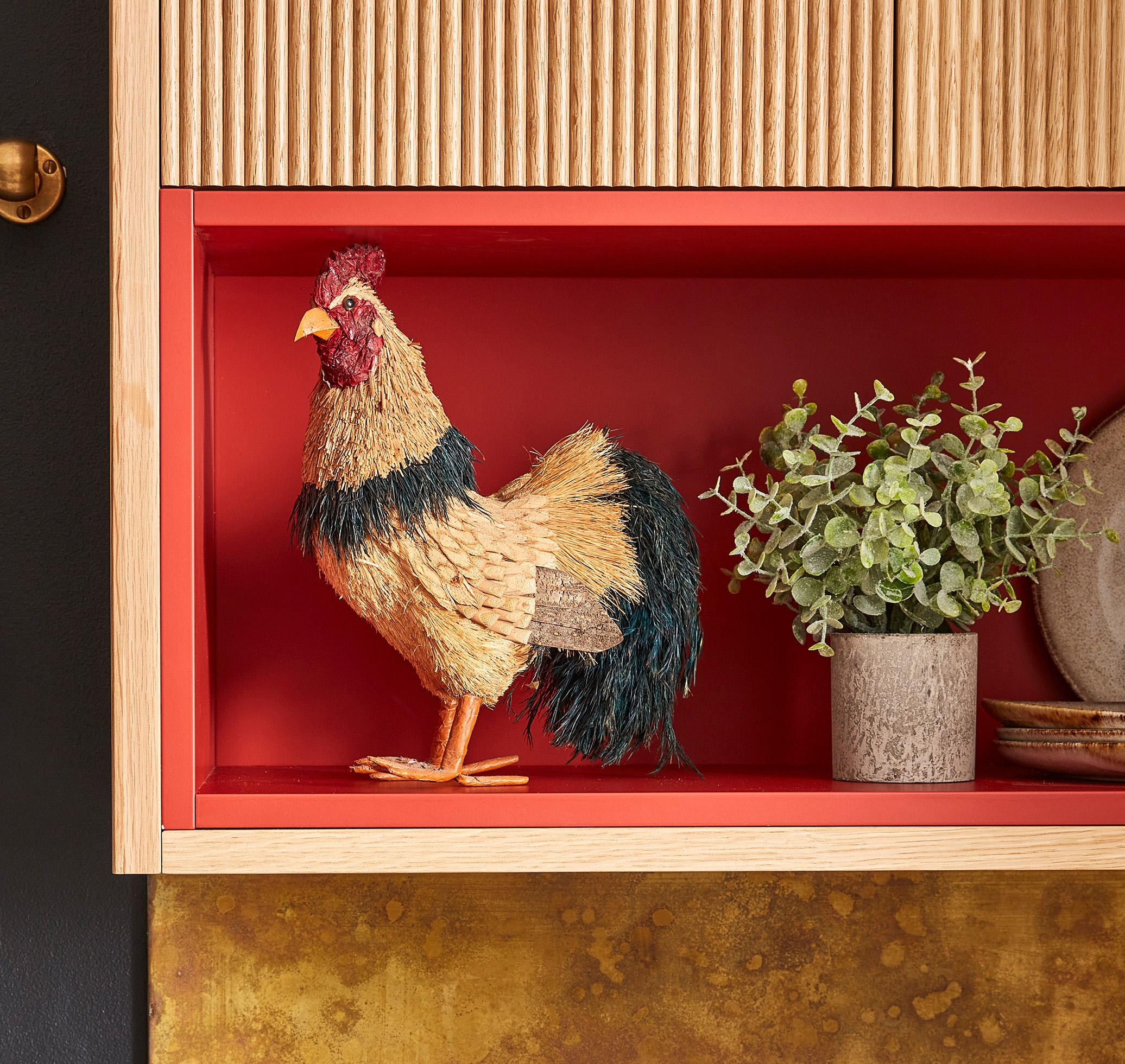 A decorative rooster figurine made of straw stands on a red shelf, next to a potted plant and a plate. The shelf is surrounded by wood and brass accents.