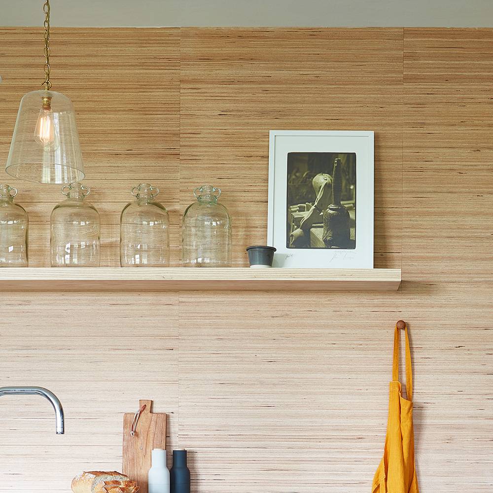 A wooden kitchen shelf holds four clear glass jars, a framed black-and-white photo, and a small cup. Below, an apron hangs on a hook and a breadboard with bread sits near a sink. A pendant light hangs above.