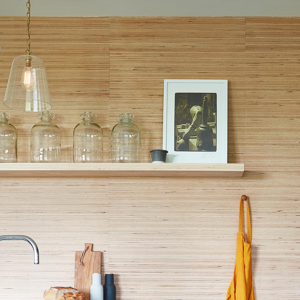 A wooden kitchen shelf holds four clear glass jars, a framed black-and-white photo, and a small cup. Below, an apron hangs on a hook and a breadboard with bread sits near a sink. A pendant light hangs above.