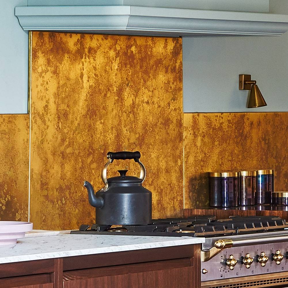A modern kitchen features a gold metallic backsplash, a black kettle on a stovetop, marble countertop, and metallic canisters. A brass wall sconce and wood cabinetry are also visible.