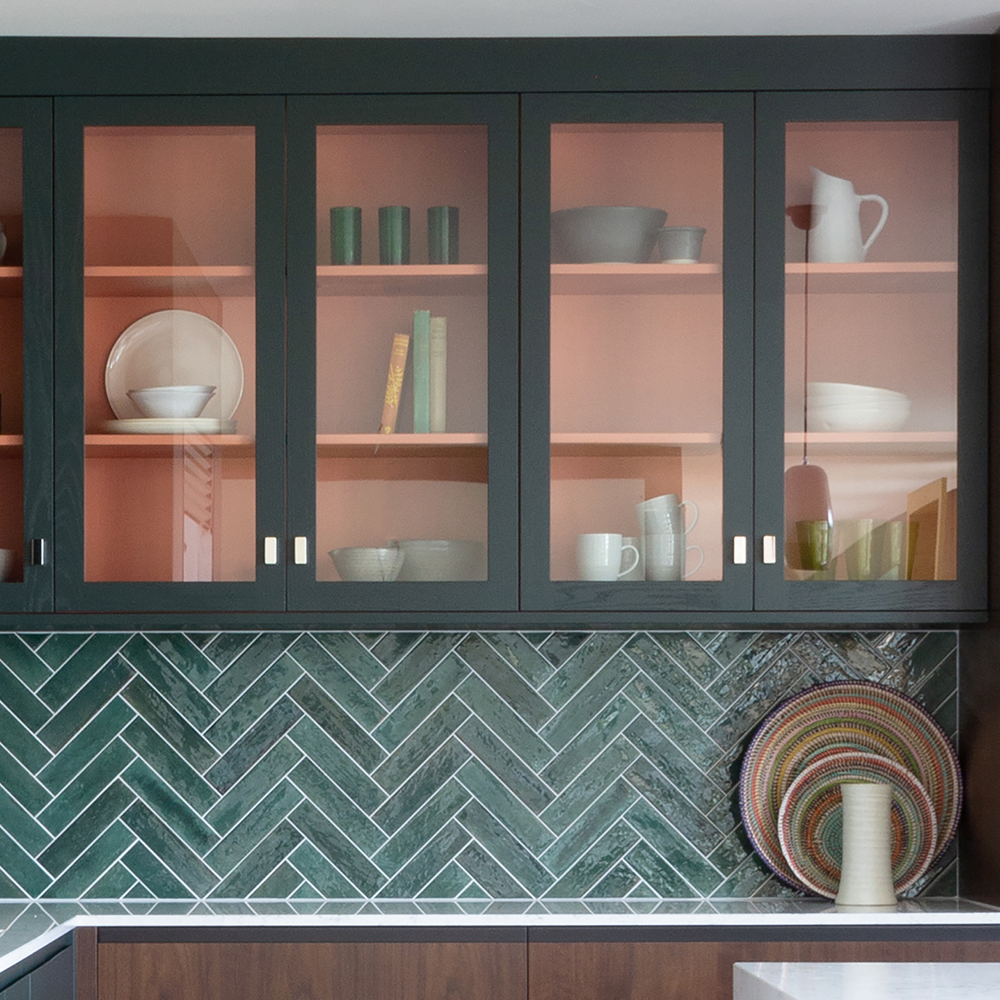 Glass-front kitchen cabinets with peach-colored interiors display dishes, cups, and books. The backsplash is dark green tile in a herringbone pattern. A colorful woven tray and a ceramic vase sit on the countertop.