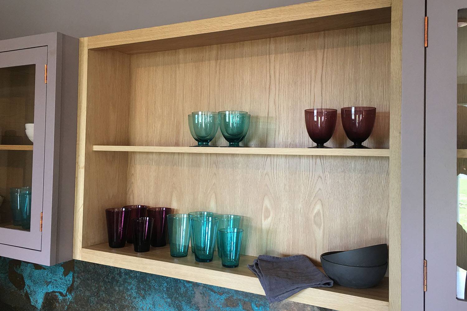 Wooden kitchen shelf with neatly arranged glassware, including teal and purple glasses, maroon goblets, and a gray bowl. Two folded gray napkins sit on the lower shelf. Brown cabinets with glass doors frame the shelf.