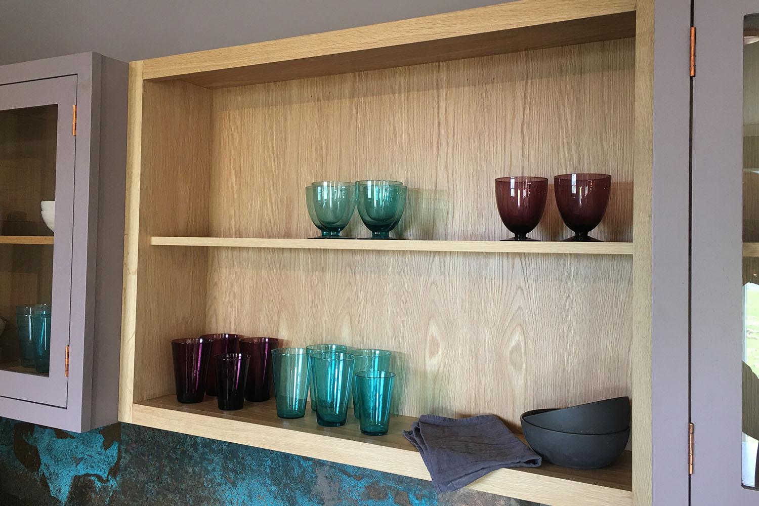 Wooden kitchen shelf with neatly arranged glassware, including teal and purple glasses, maroon goblets, and a gray bowl. Two folded gray napkins sit on the lower shelf. Brown cabinets with glass doors frame the shelf.