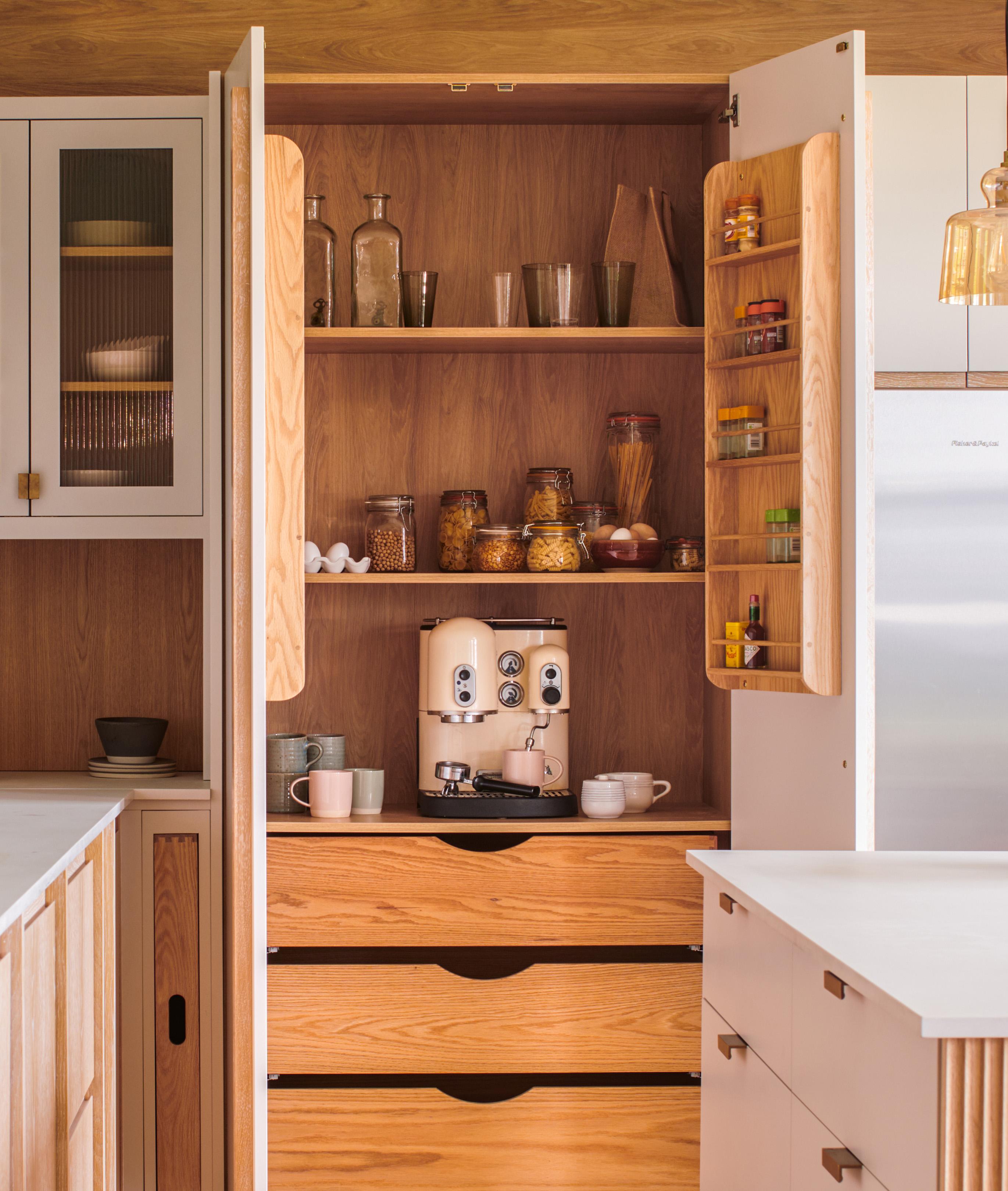 Open kitchen cabinet with wooden shelves holding jars, bottles, and a coffee machine. Three drawers below and smaller shelves on both doors contain spices. Warm lighting and modern design elements are present.