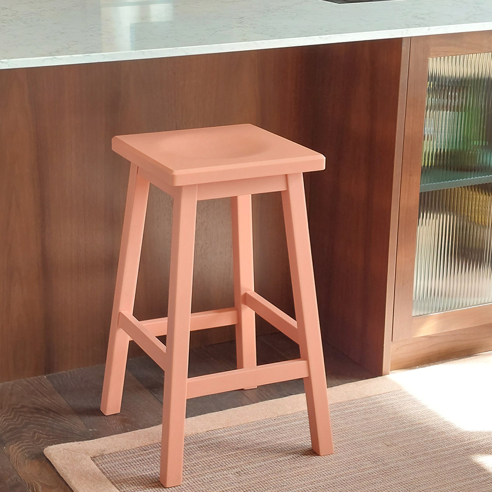 A peach-colored wooden stool with a square seat stands on a rug beside a kitchen counter with a marble top and a wood-paneled cabinet.