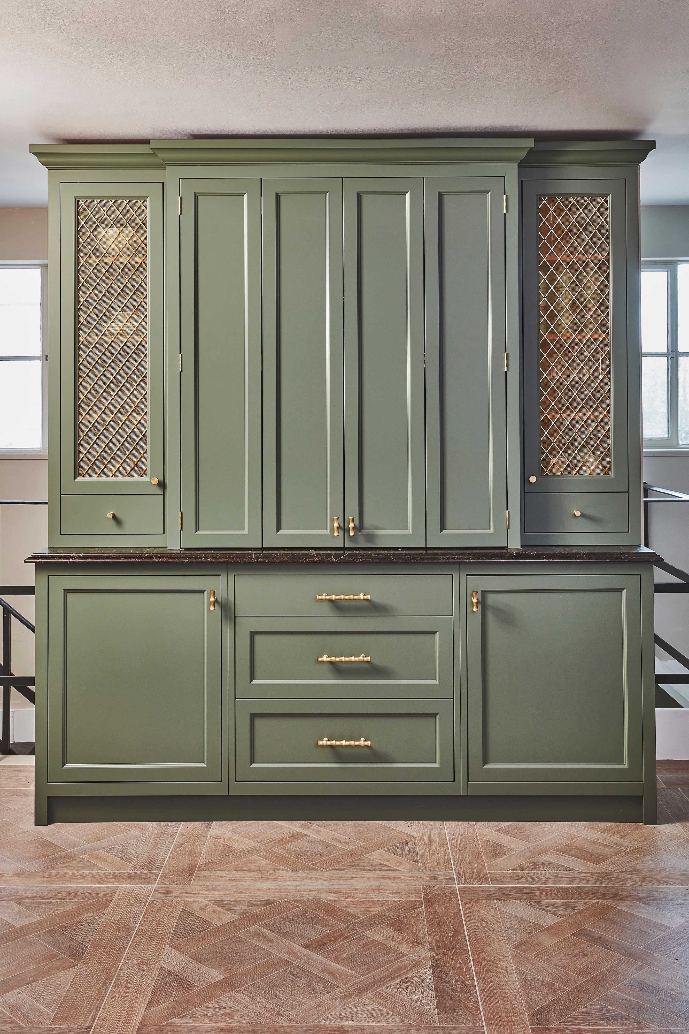 Sage green kitchen cabinets with gold mesh panel inserts, brass handles, and a dark countertop, set against a neutral wall with two windows in the background and geometric patterned wood flooring.