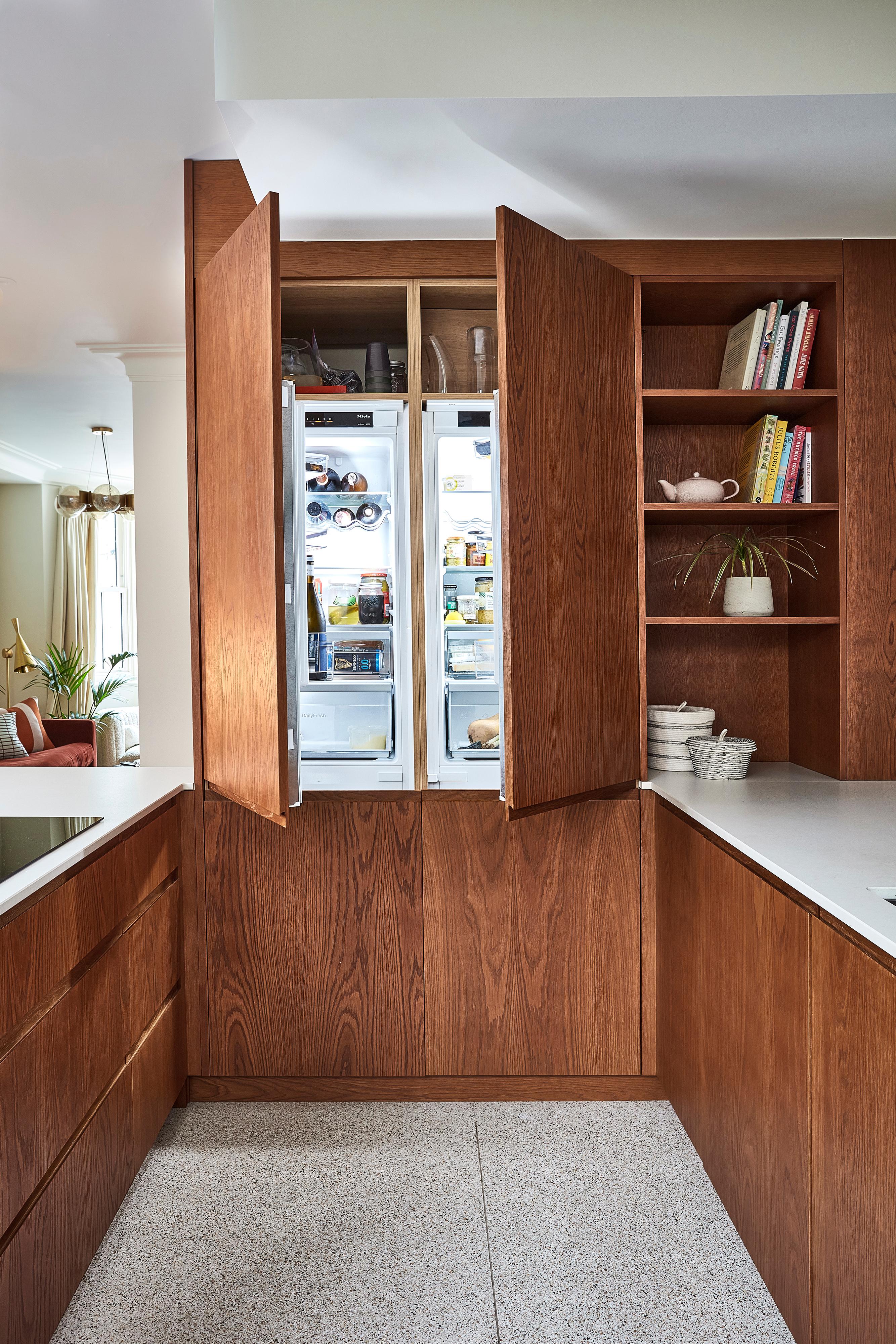 Modern kitchen with sleek wooden cabinets and white countertops. Open shelving displays books and a plant. Natural light streams in through a window, creating a warm, inviting atmosphere.