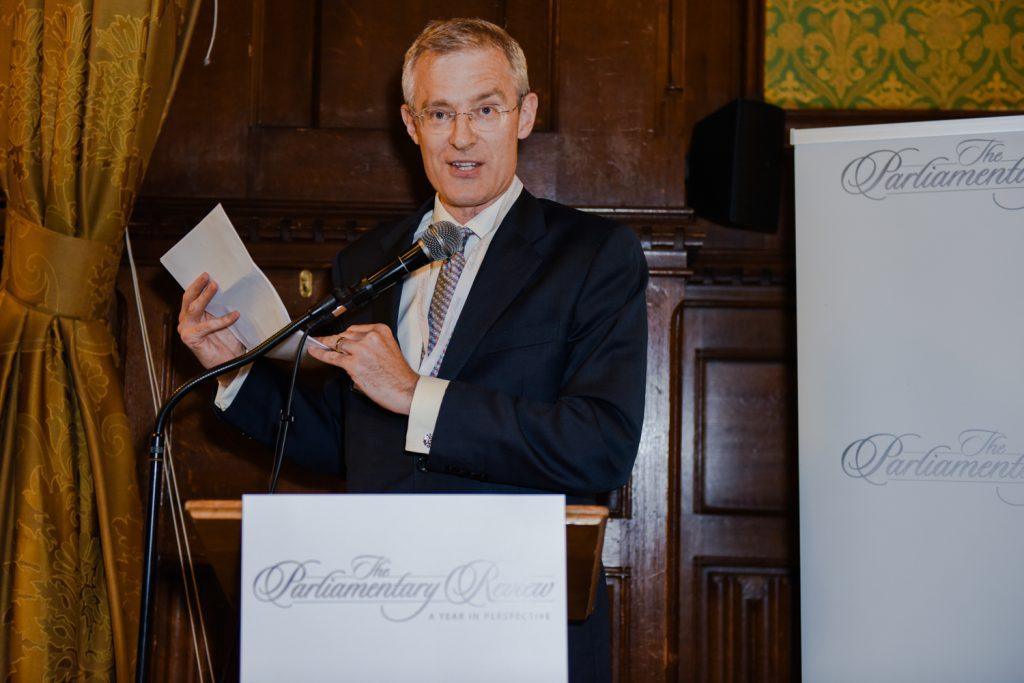 A man with glasses and a dark suit holds notes while speaking into a microphone at a "The Parliamentary Review" podium.
