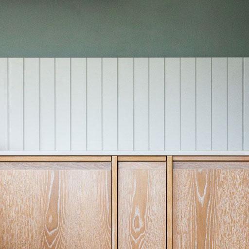 Wooden cabinets with a light, natural finish are set against a wall with vertical white paneling, topped by a gray-green painted section. The scene conveys a minimalist, modern design aesthetic.