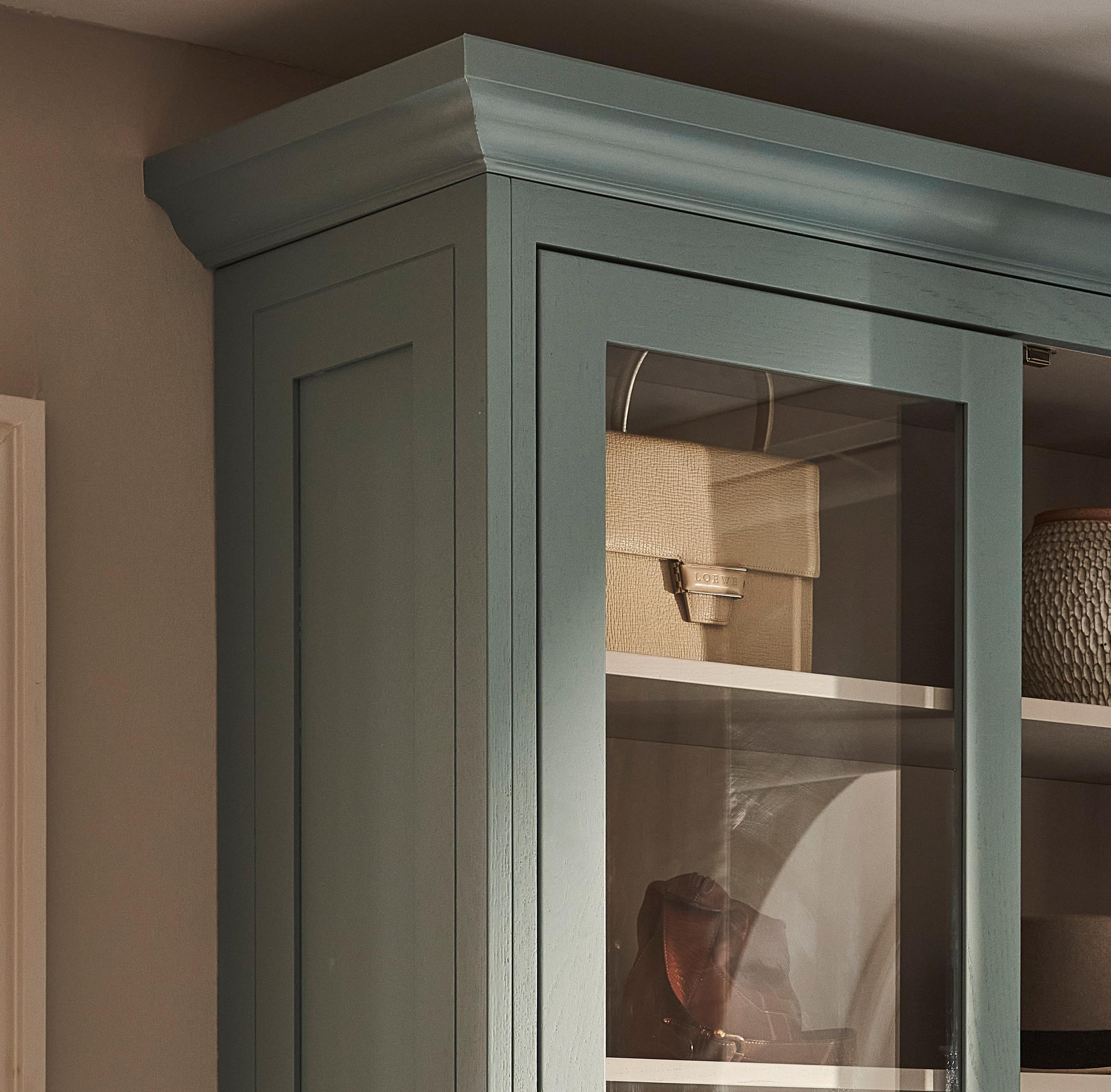 A close-up of the top corner of a blue-green glass-front cabinet with crown molding, showing neatly arranged items on the shelves inside, including a beige box and a textured container.