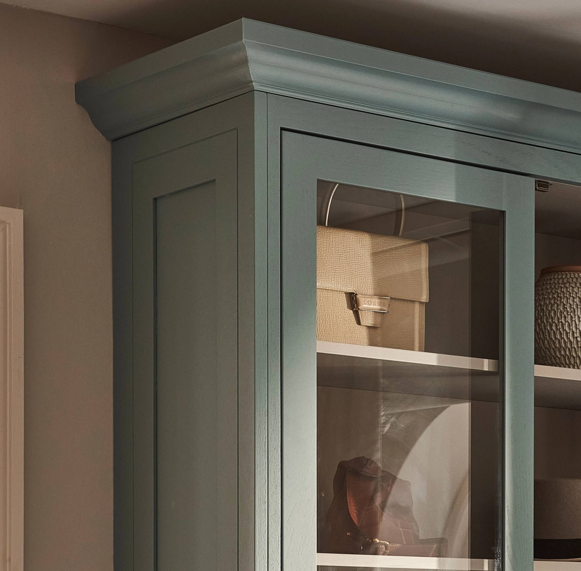A close-up of the top corner of a blue-green glass-front cabinet with crown molding, showing neatly arranged items on the shelves inside, including a beige box and a textured container.